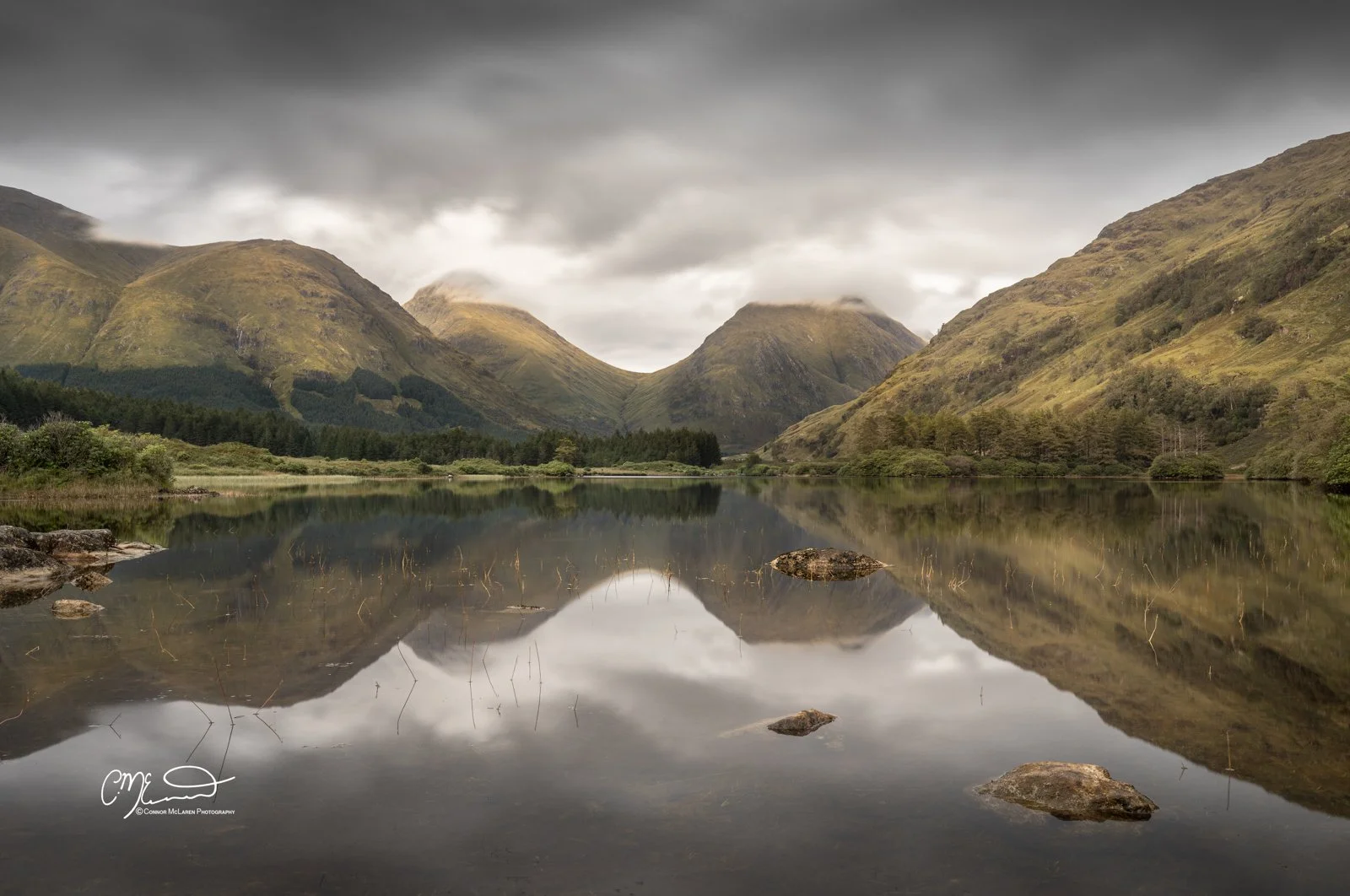 Lochan Urr, Glen Coe and Glen Etive, Western Highlands, Scotland. Calm Loch surrounded by green hills and mountains, overcast sky with reflections in the water, rocks in the foreground.