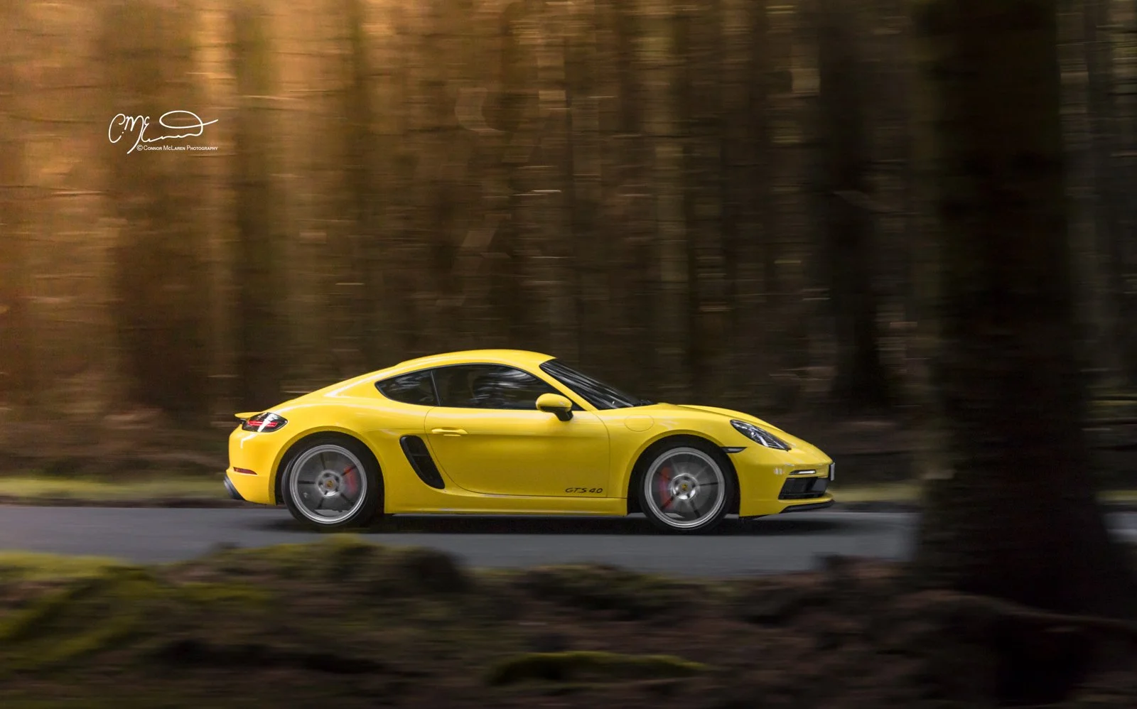 Yellow Porsche 718 Cayman GTS Sports car driving on a tree-lined road with blurred background.