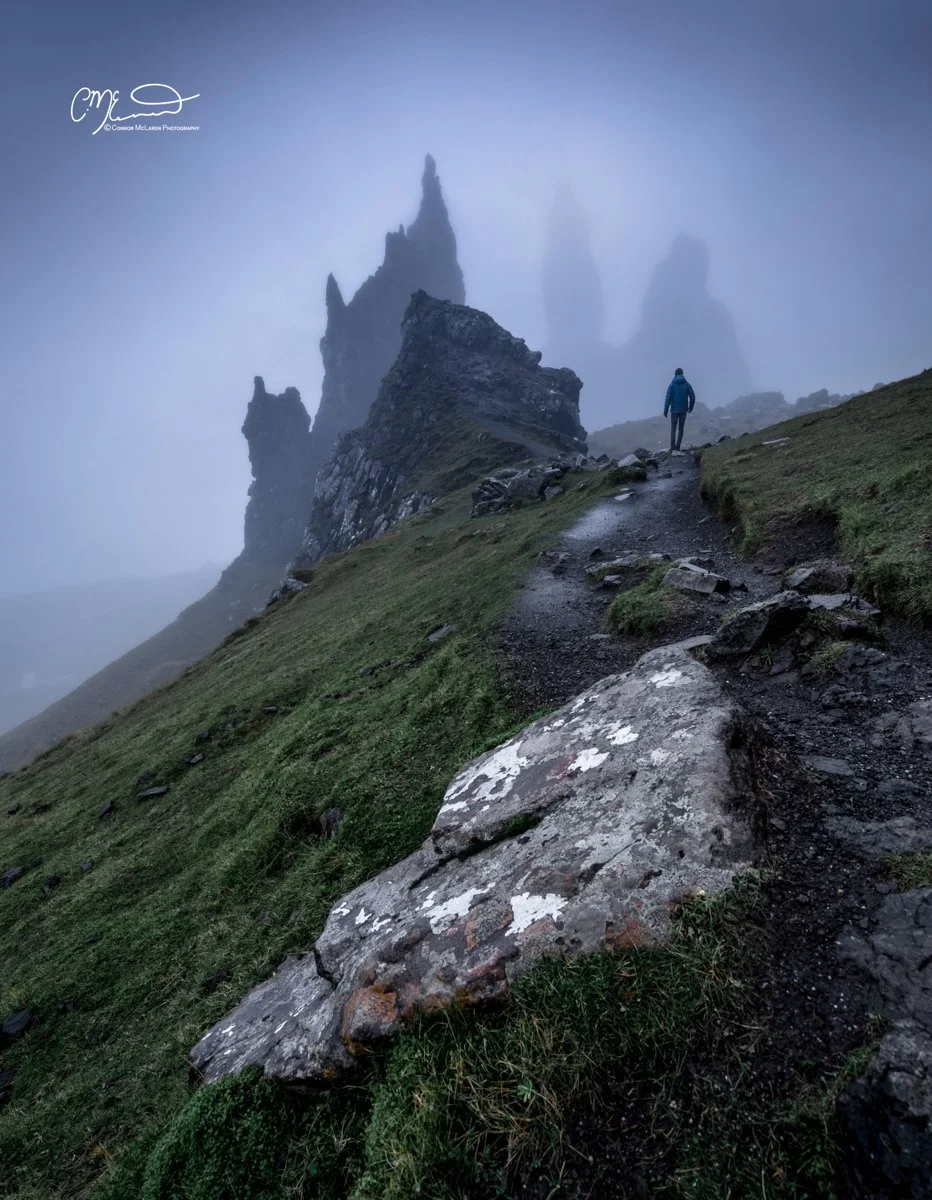 Scotland, Isle of Skye, Old Man of Storr. A person hiking on a narrow, muddy trail on a steep hillside covered with green grass, with tall, jagged rock formations in the foggy background.
