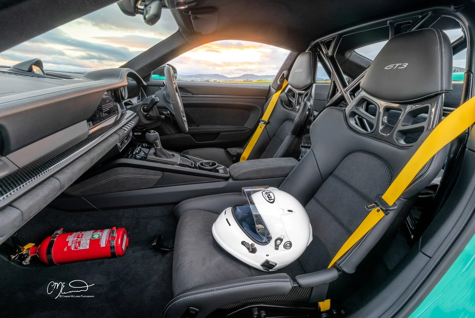 Mint Porsche 911 GT3 992. Interior of a race car with two racing seats, a white helmet on the passenger seat, a yellow racing harness, and a fire extinguisher on the floor, with a scenic landscape visible through the windshield.
