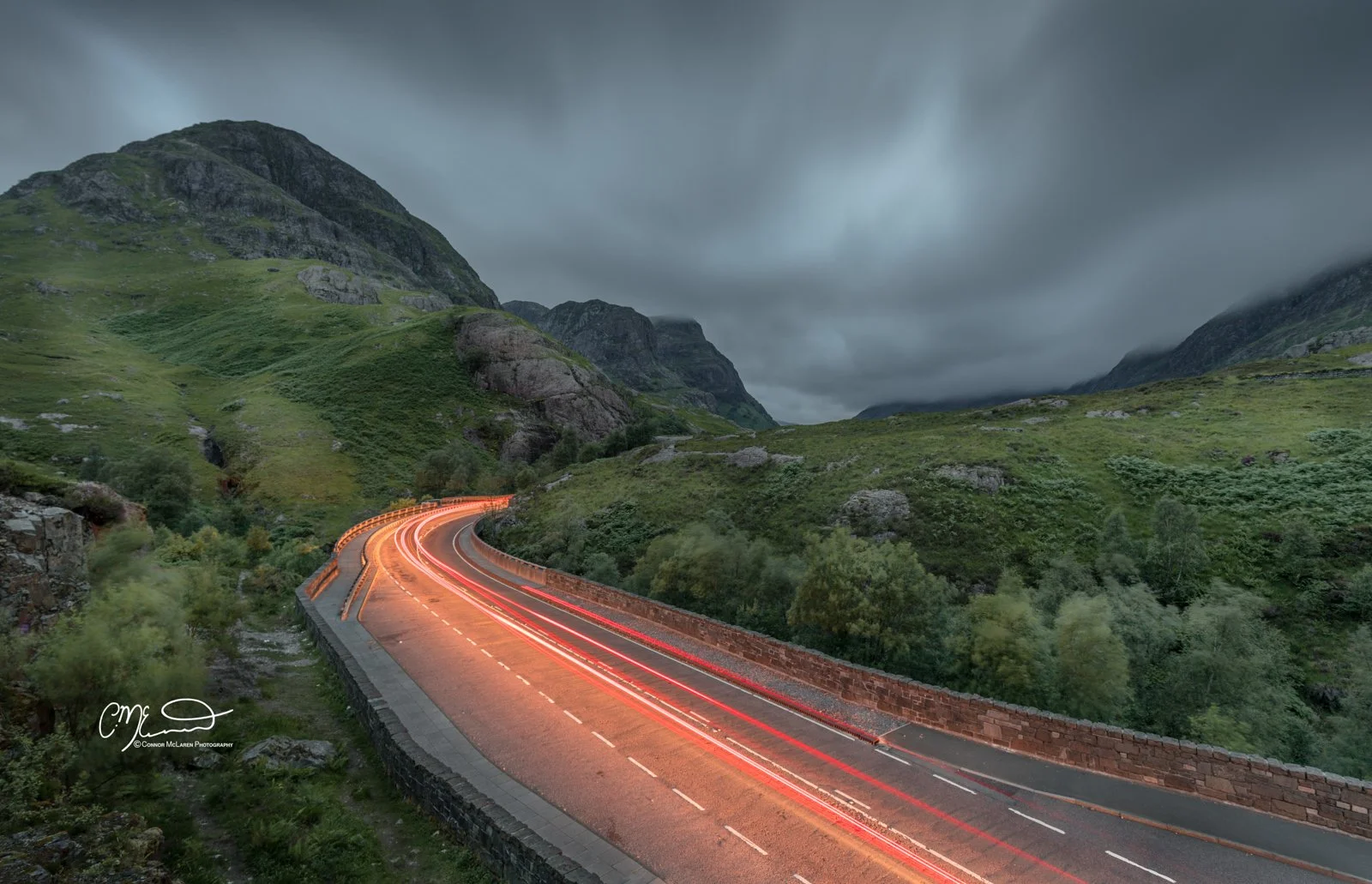 Glen Coe and Glen Etive, Western Highlands, Scotland. Curving mountain road at dusk with light trails from moving vehicles, surrounded by green hills and cloudy sky.