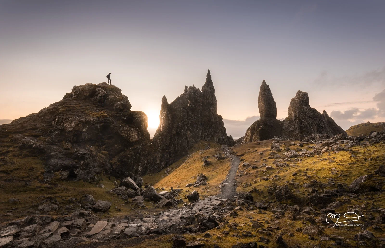 Scotland, Isle of Skye, Old Man of Storr. A hiker standing on a rocky hill at sunrise or sunset, overlooking rugged rock formations on a grassy landscape with a dirt trail in the foreground.