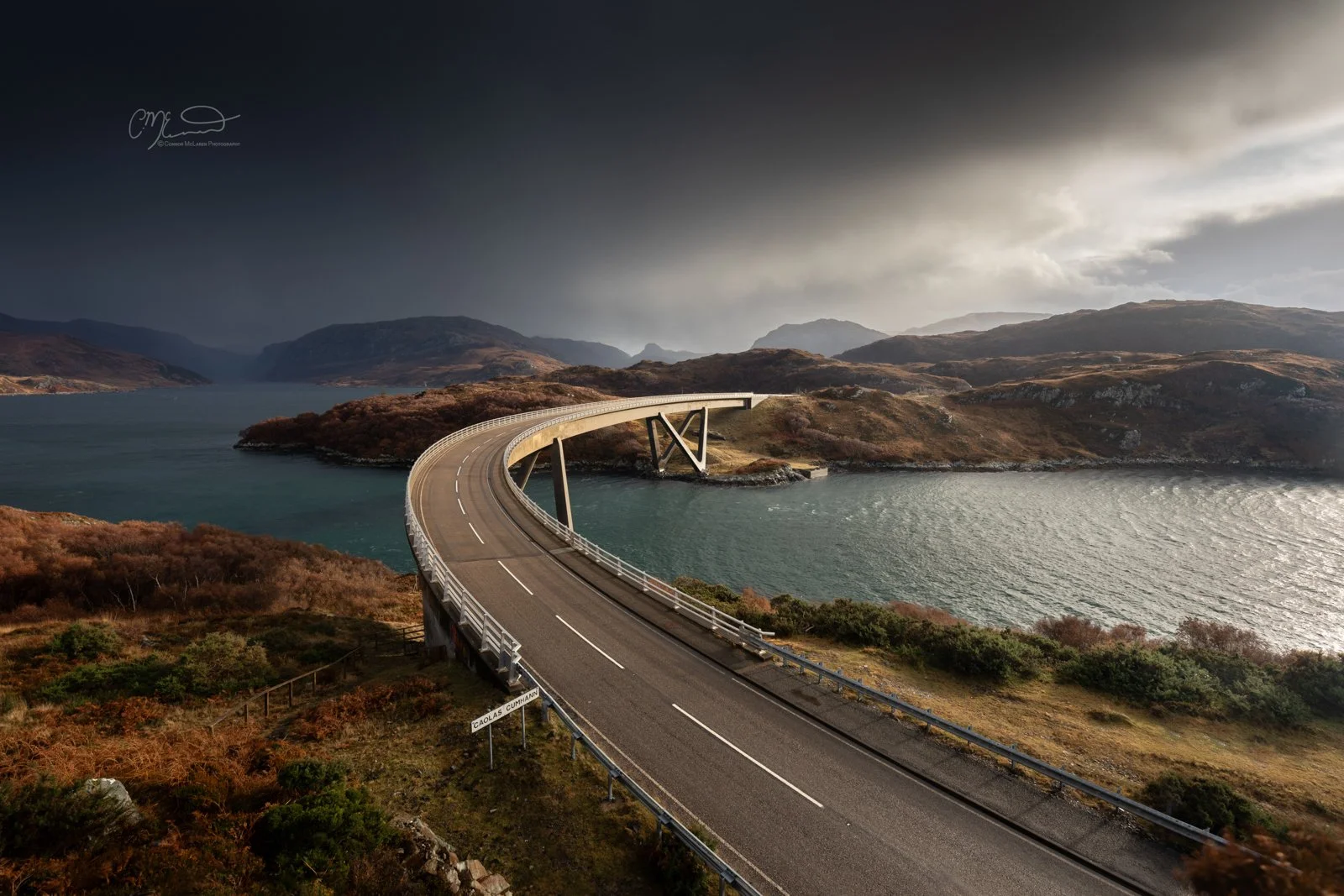 Scotland, north coast 500, NC500, Kylesku Bridge. A curved bridge extending over a body of water surrounded by hilly terrain with dark stormy clouds overhead.