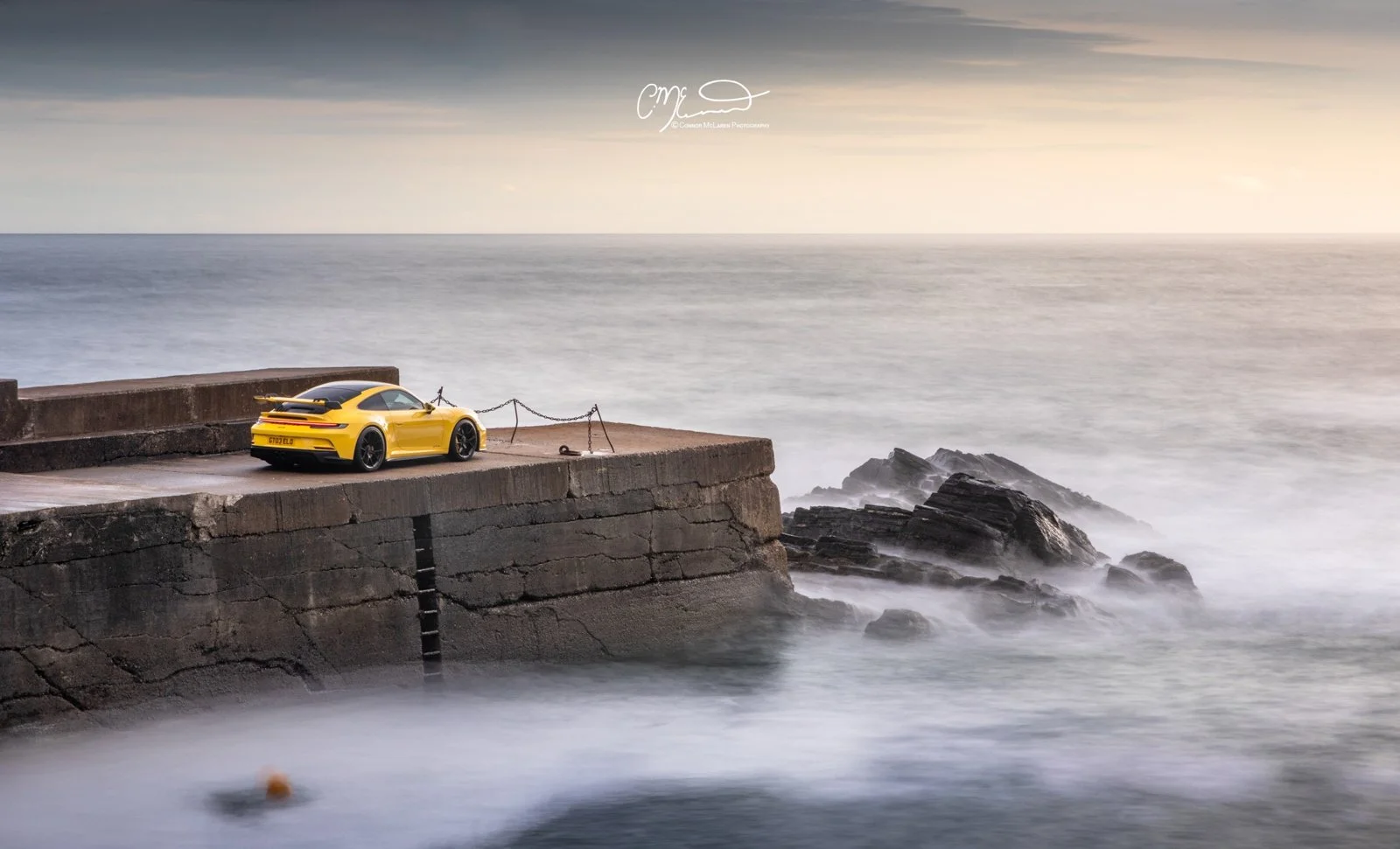 A yellow Porsche GT3 sports car parked on a concrete pier by the ocean during a cloudy day.