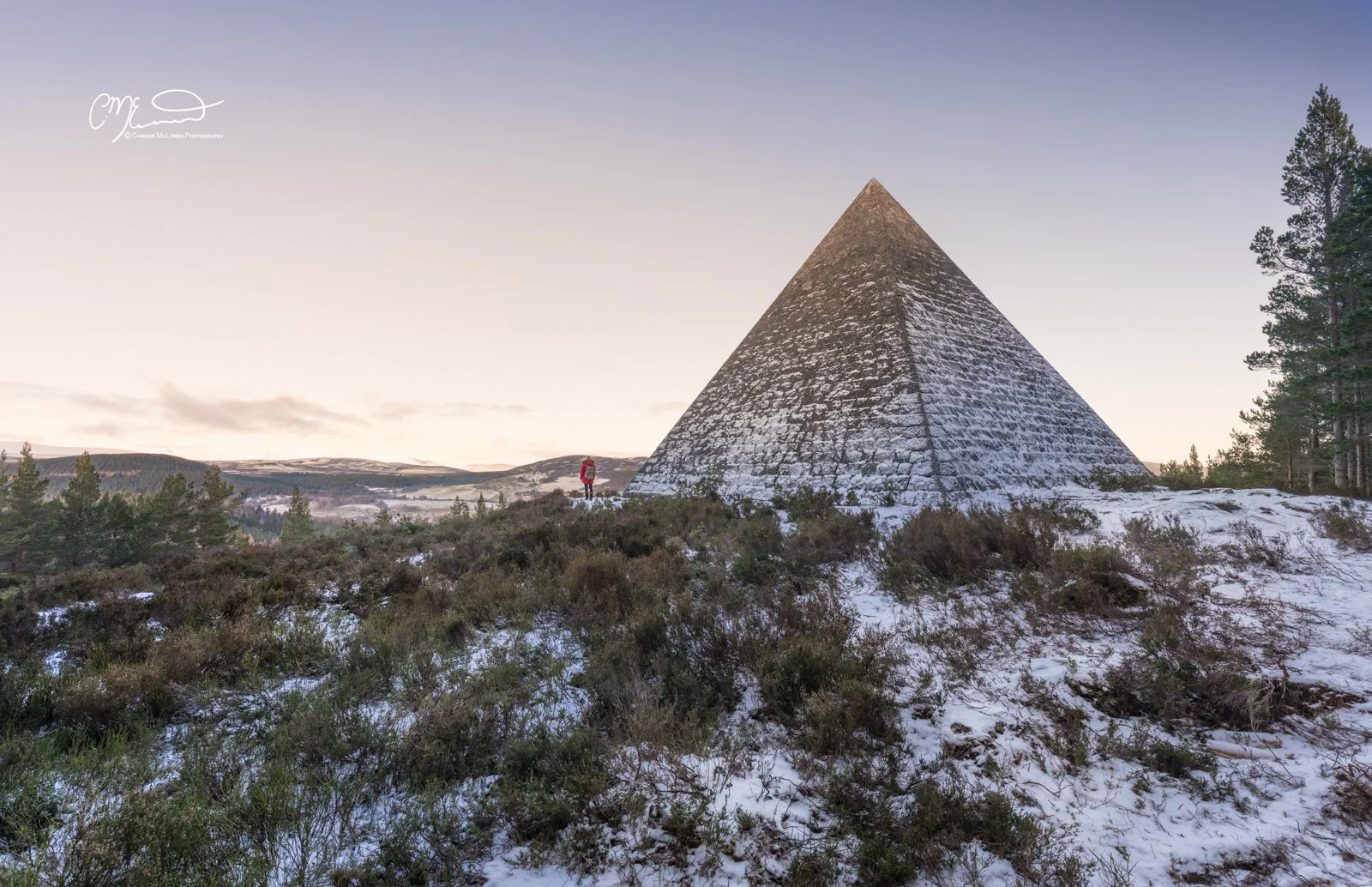 Scottish Central Highlands, Scotland, Pyramid. A person in a red jacket standing near a large stone pyramid-shaped structure in a snowy landscape with trees and distant hills at sunset or sunrise.