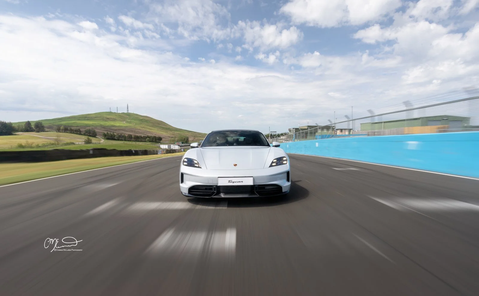 A white Porsche Taycan sports car driving on a race track with green hills, a cloudy sky, and a blue barrier wall on the side.