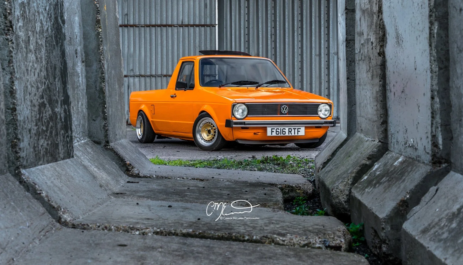 Orange vintage Volkswagen Caddy pickup truck parked between concrete barriers and corrugated metal fencing.