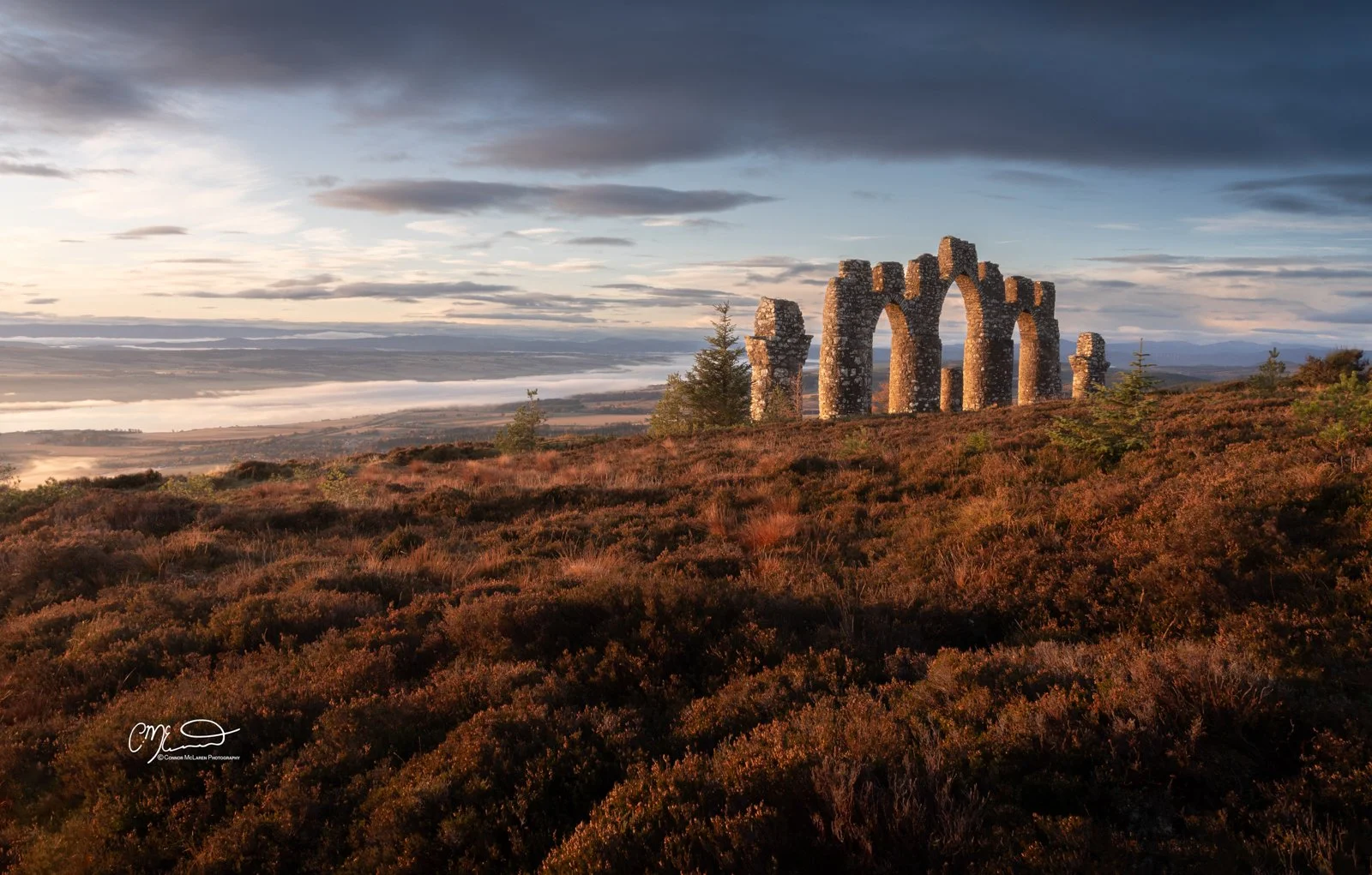Scotland, north coast 500, NC500, Fyrish Monument. Ancient stone ruins with arches on a hilltop at sunrise, overlooking a landscape with fog over fields and a river.