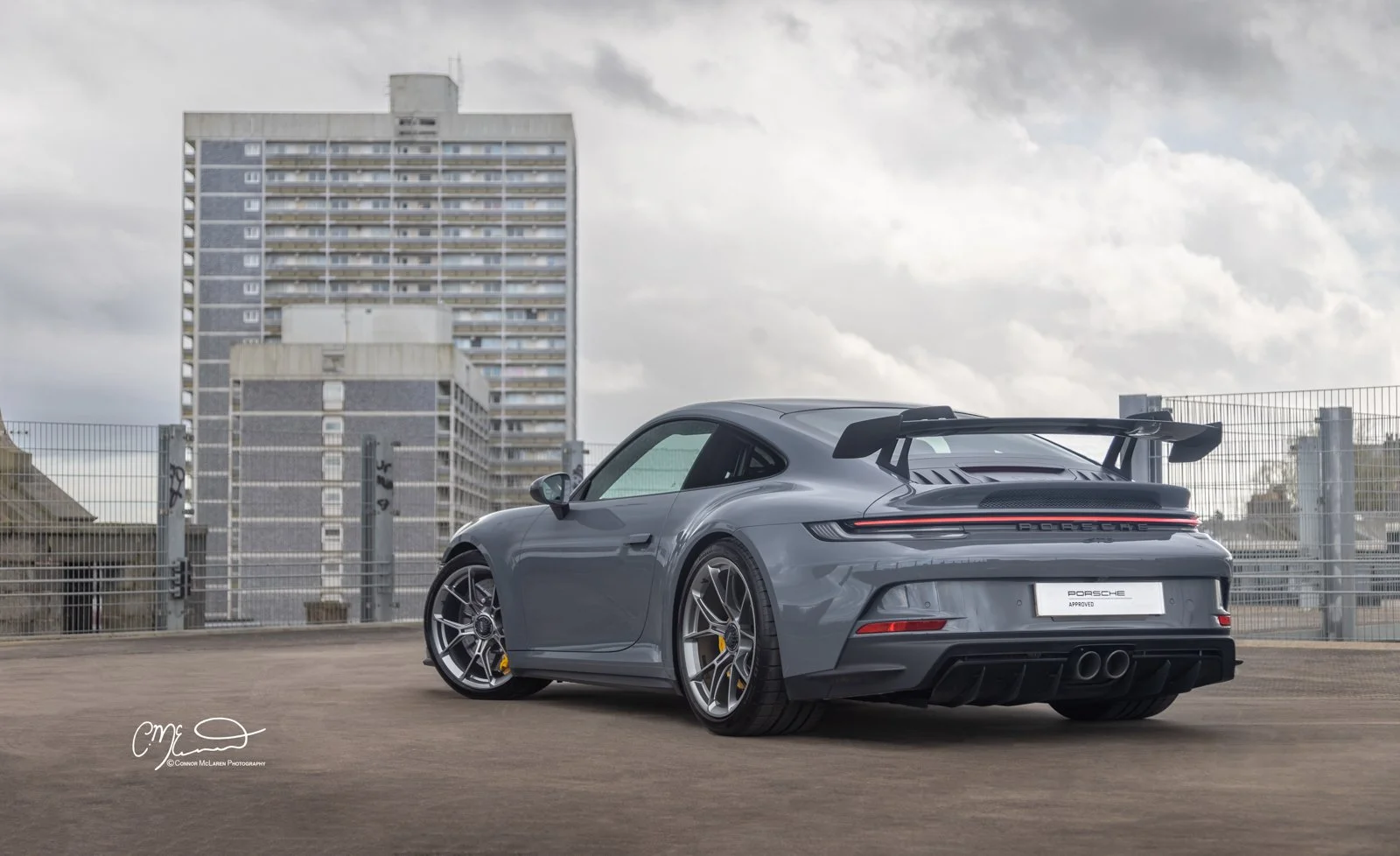 Gray Porsche 911 GT3 sports car parked on rooftop with city buildings and cloudy sky in background.