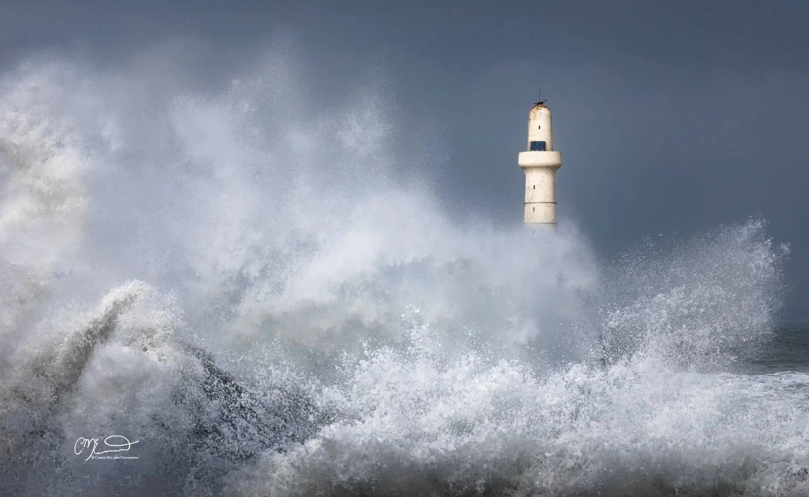Aberdeen lighthouse, storm, North East Scotland. A lighthouse on a rocky coast with large waves crashing against the rocks, creating spray and foam, under a cloudy sky.