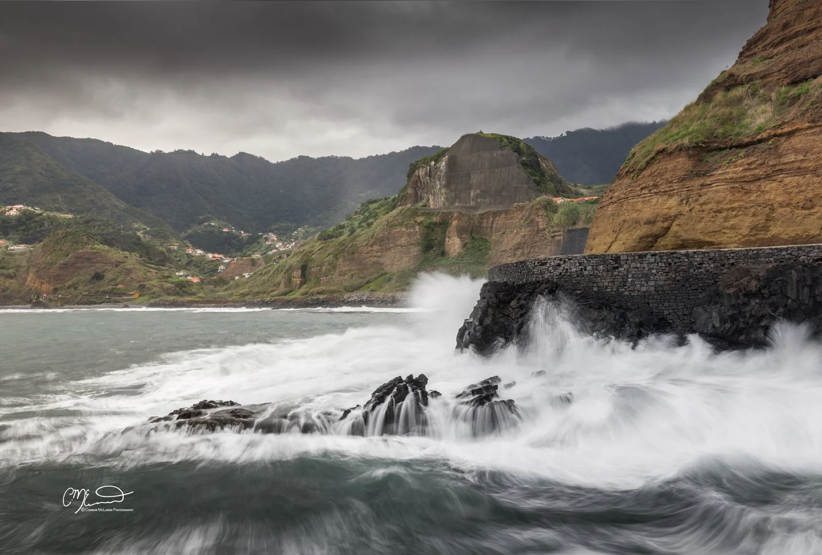 Volcanic rock, porto da cruz, Madeira Portugal. Stormy ocean waves crashing against rocky shoreline with green cliffs and hills in the background under dark cloudy sky.