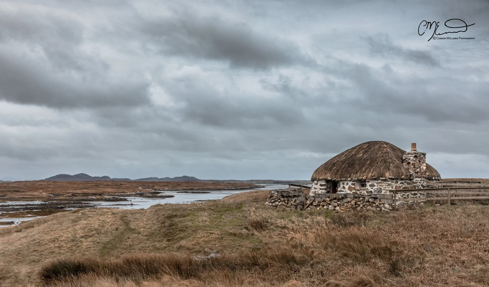 Scotland, Lewis Harris & Outer Hebrides, South Uist. A stone cottage with a thatched roof situated near a body of water under a cloudy sky, surrounded by grassy terrain on a cloudy day.
