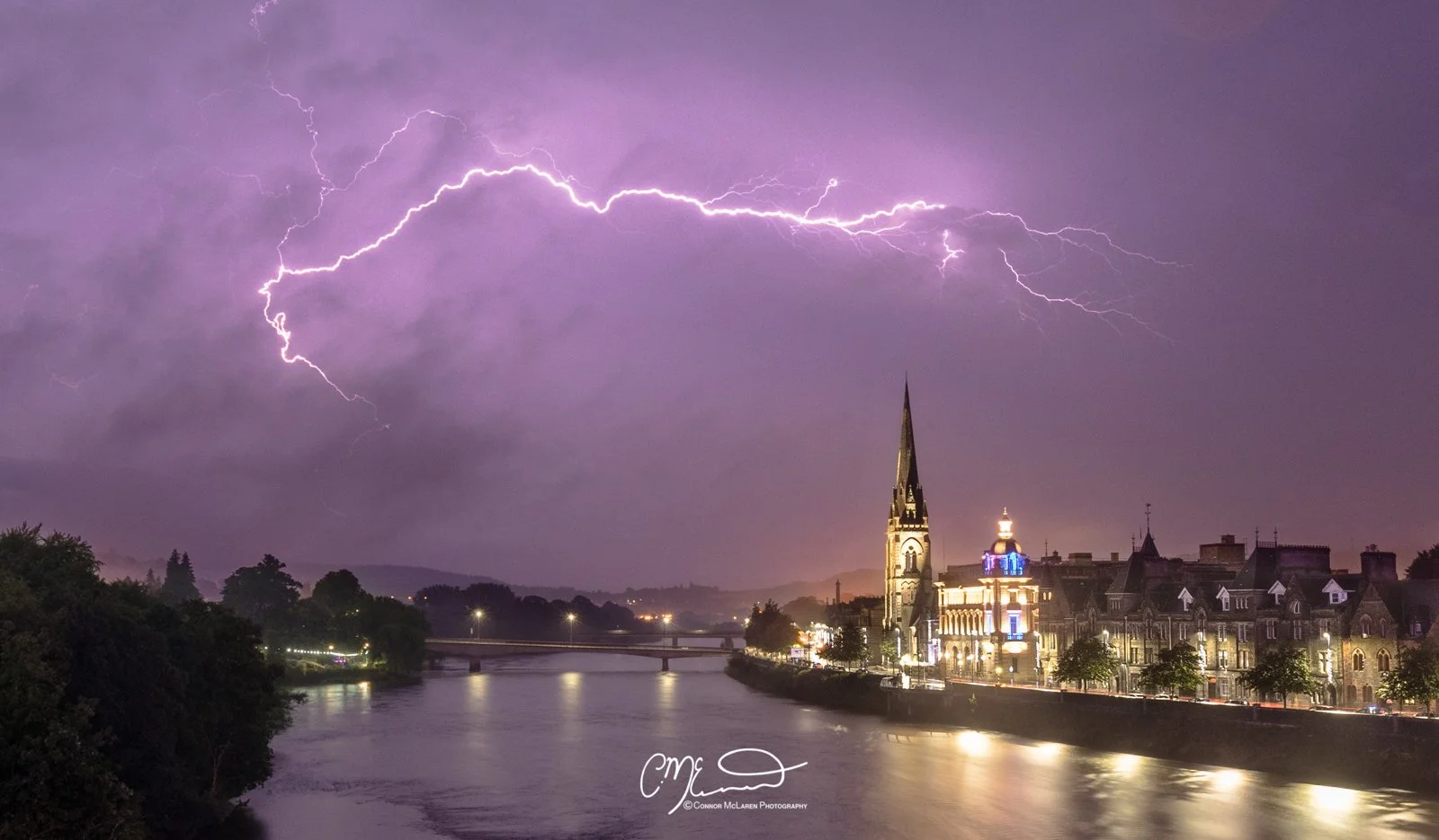 Perth Scotland Lightning storm. Nighttime scene of a city with a river in the foreground, illuminated historic buildings including a church with a tall spire, and a lightning storm with bright purple lightning bolts in the cloudy sky.