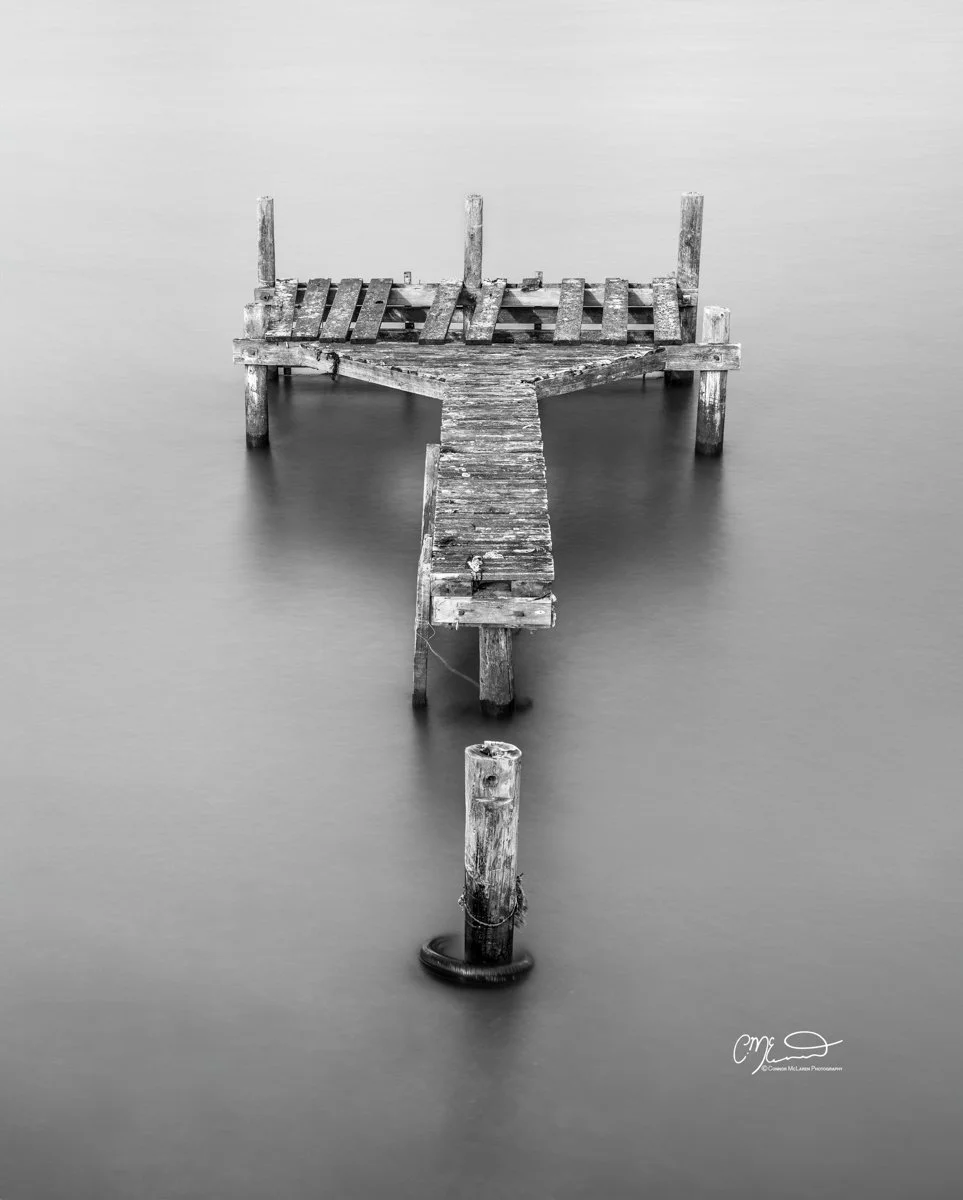 Scotland, abandoned pier. Black and white photo of an old, weathered wooden dock extending into calm water, with some of the dock's platforms broken or missing.