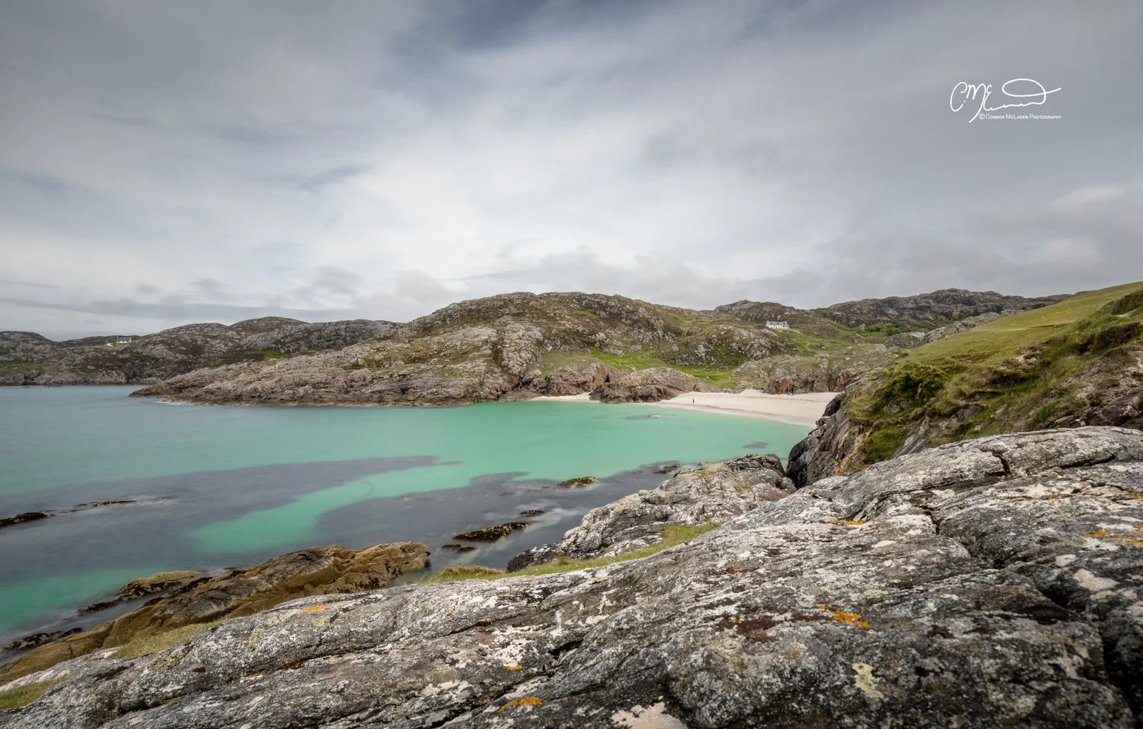 Scotland, north coast 500, NC500, Achmelvich beach. A coastal landscape with rocky foreground leading to a turquoise bay and beach, surrounded by green hills and cloudy sky.