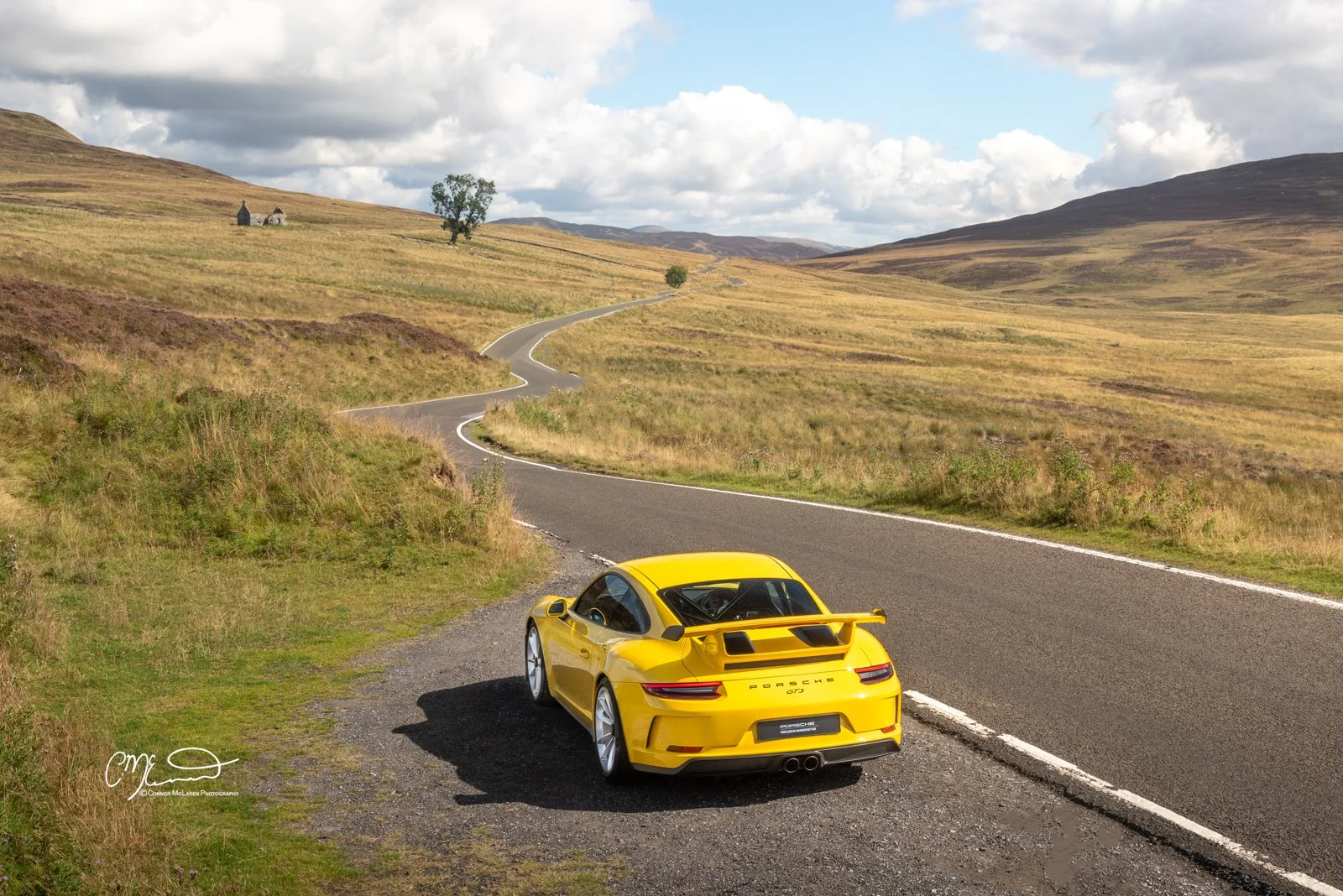 A yellow Porsche 911 GT3 sports car parked on the side of a winding country road surrounded by grassy hills under a partly cloudy sky.