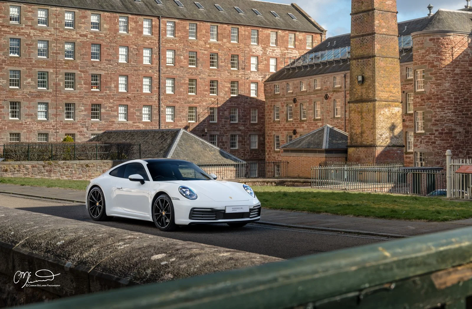 A white Porsche 911 sports car parked on a city street with brick historical buildings in the background.