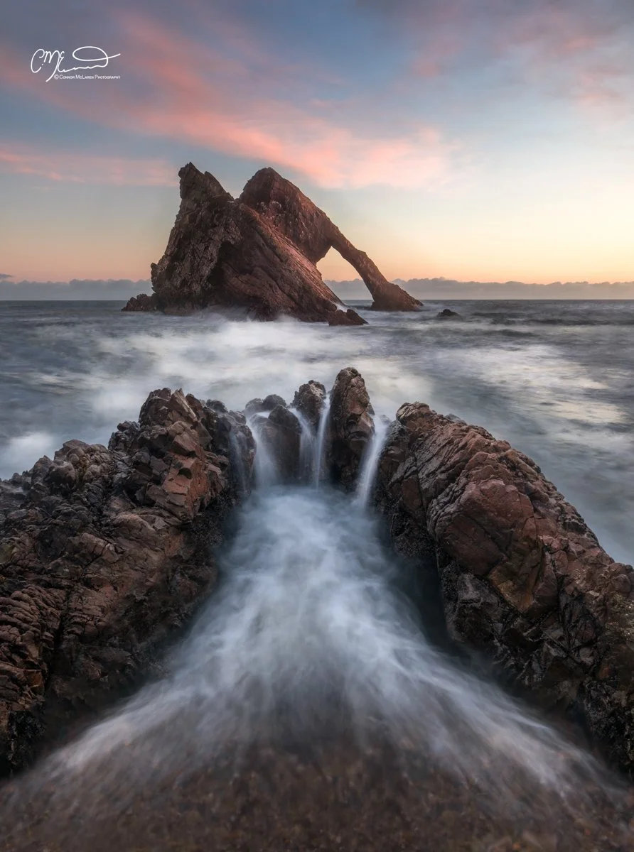 Bucket list locations, Scotland, Bow Fiddle Rock. Ocean waves crashing against rocky shore with a large, arch-shaped rock formation in the distance at sunset, colorful sky with pink and orange hues.