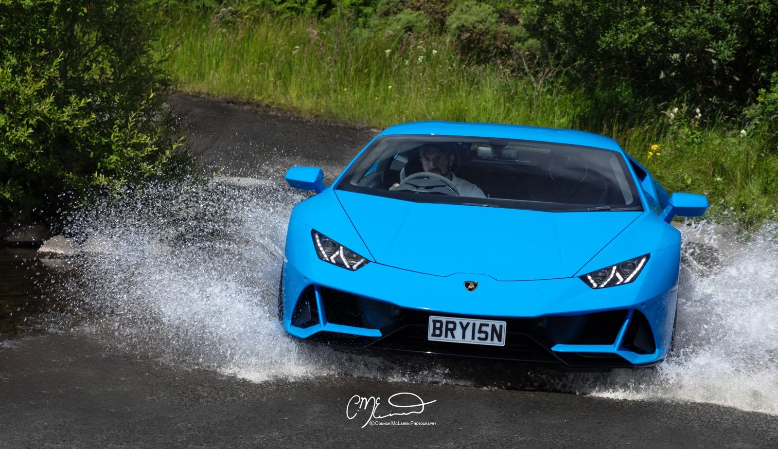 A blue Lamborghini huracan evo sports car driving on a wet road, splashing water, with greenery and trees in the background.