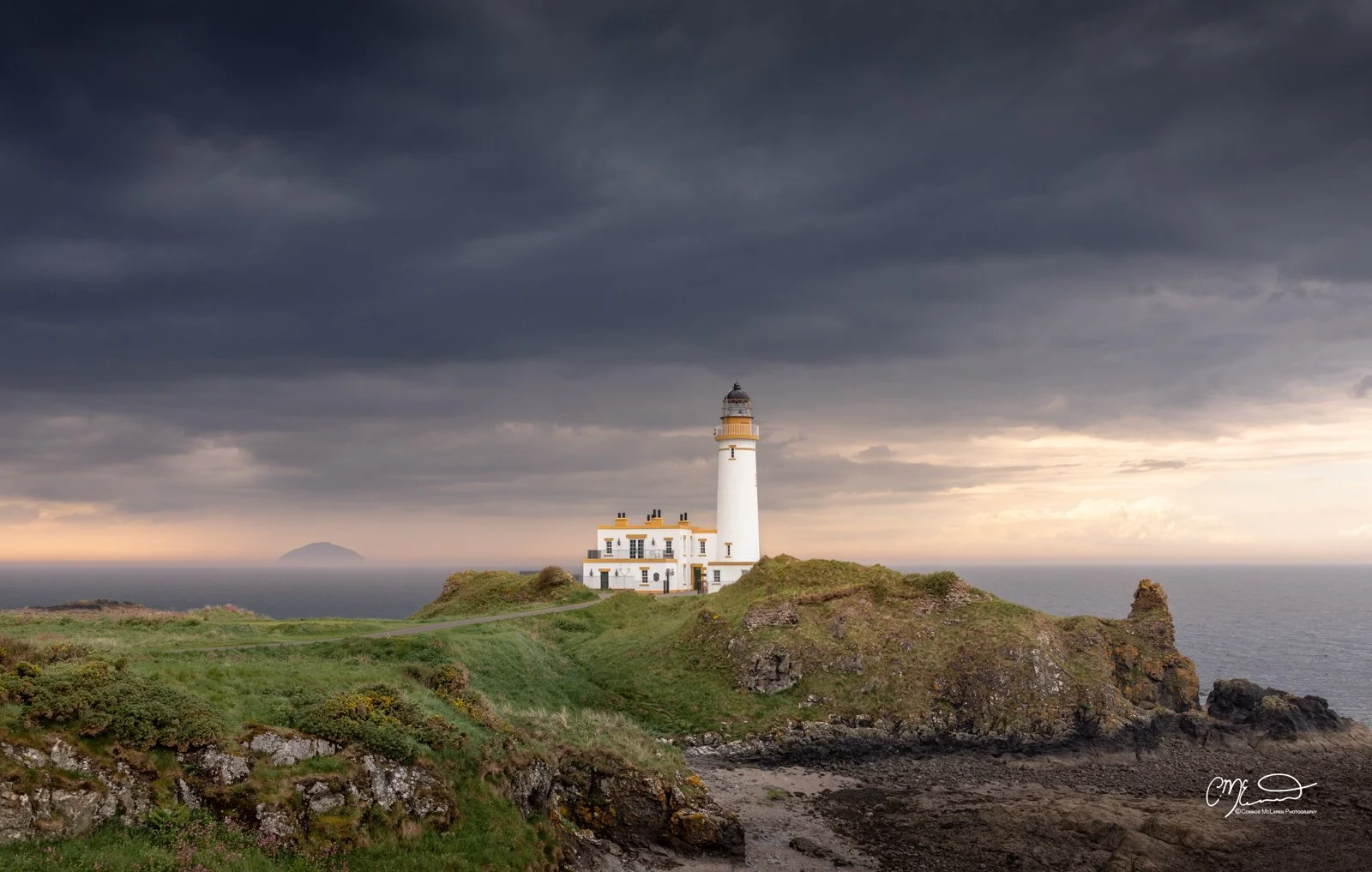 Scottish Lowlands, Scotland, Turnberry. A lighthouse on a grassy cliff overlooking the ocean under a dark, cloudy sky at sunset.
