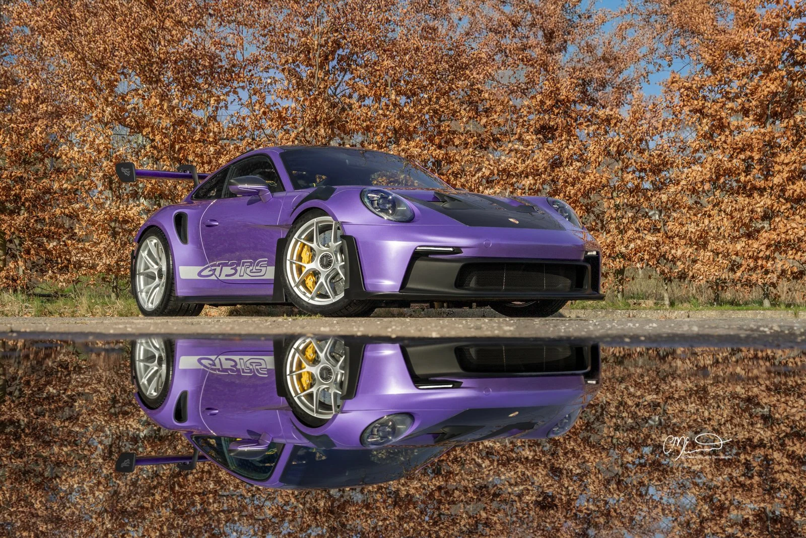 Porsche 911 GT3 RS in Paint to sample purple, weissach. A purple Porsche sports car with black accents and yellow brake calipers, reflected in a puddle on the ground, with a background of orange and brown autumn leaves.