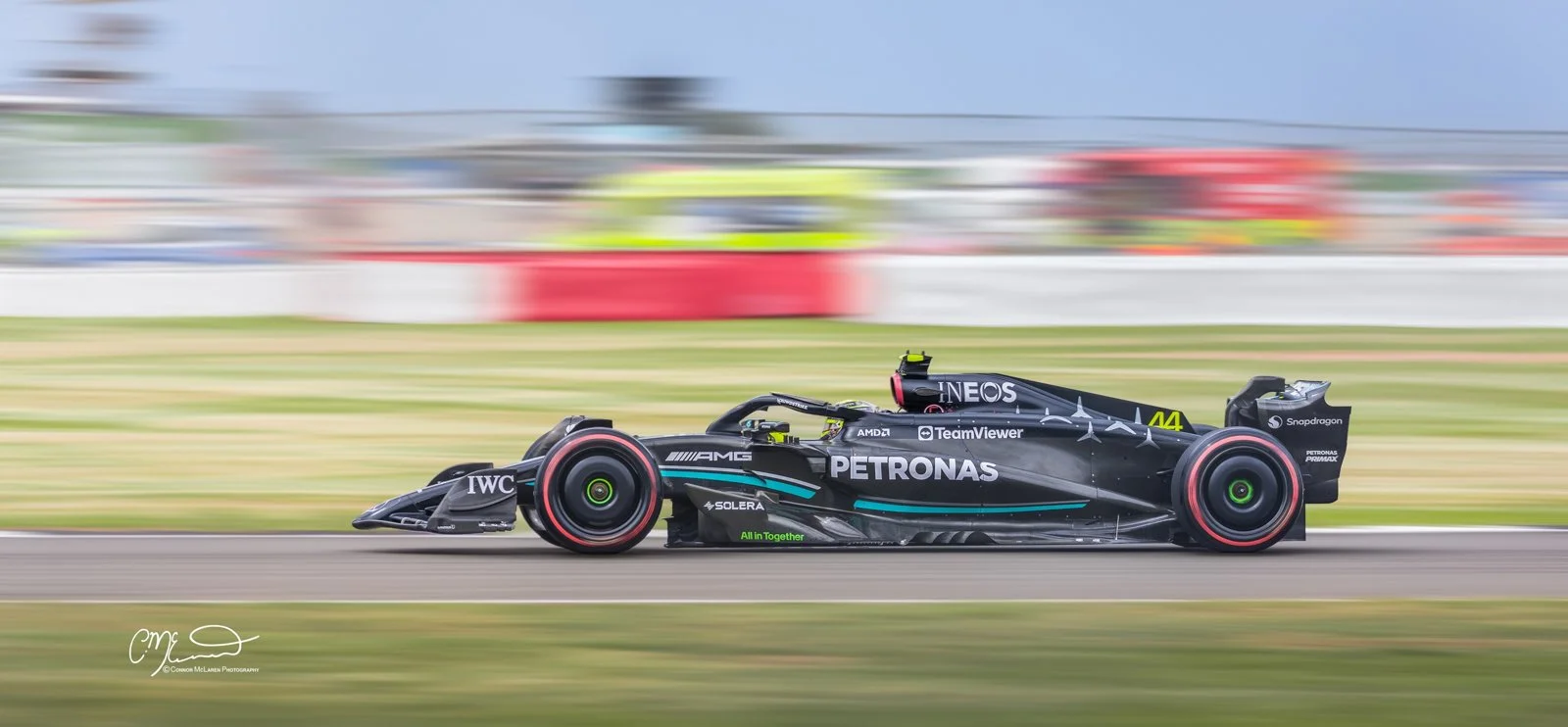 Mercedes Formula One car driver by lewis Hamilton at Silverstone. A sleek black Mercedes-AMG F1 race car on a racetrack, with vibrant sponsors and logos, in motion blur as it speeds past, with blurred spectators and track barriers in the background.