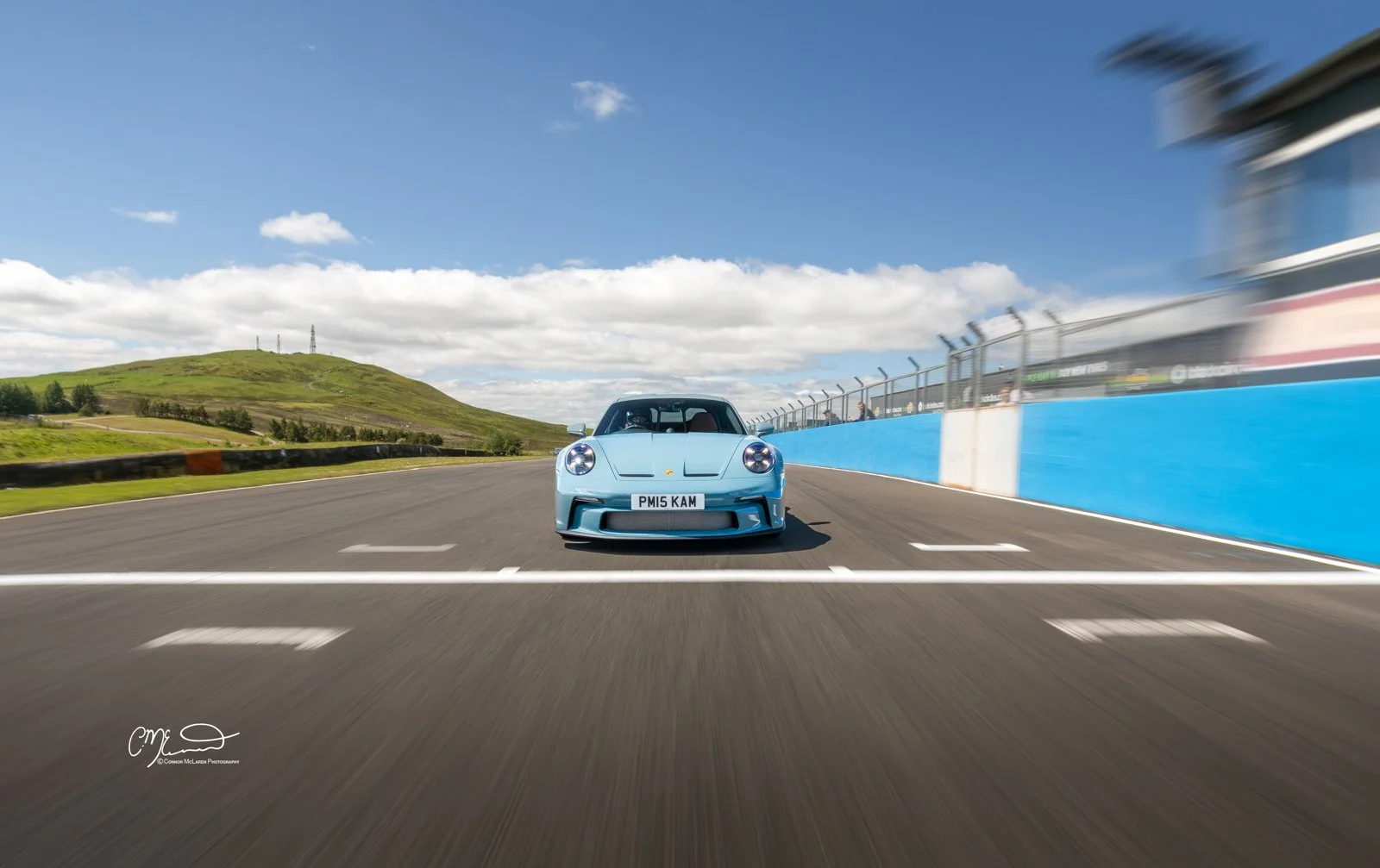 Porsche 911 ST on track at Knockhill. A blue sports car driving on a race track with a blue wall on the right and a scenic hillside with green grass and trees in the background under a partly cloudy sky.