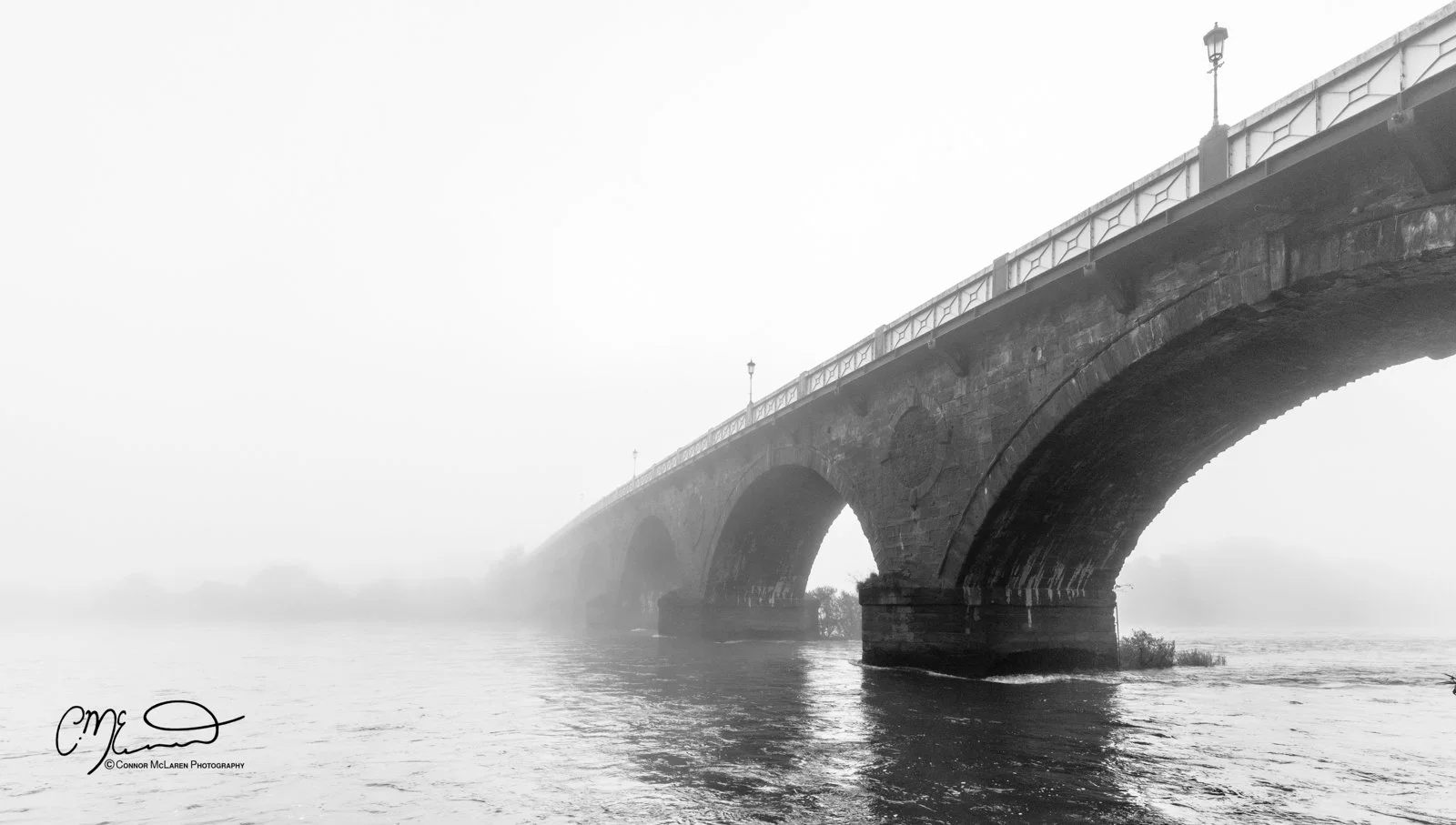 Scotland, River Tay Perth. A black and white photo of a bridge arching over a river, with fog obscuring the background and a hazy sky above.