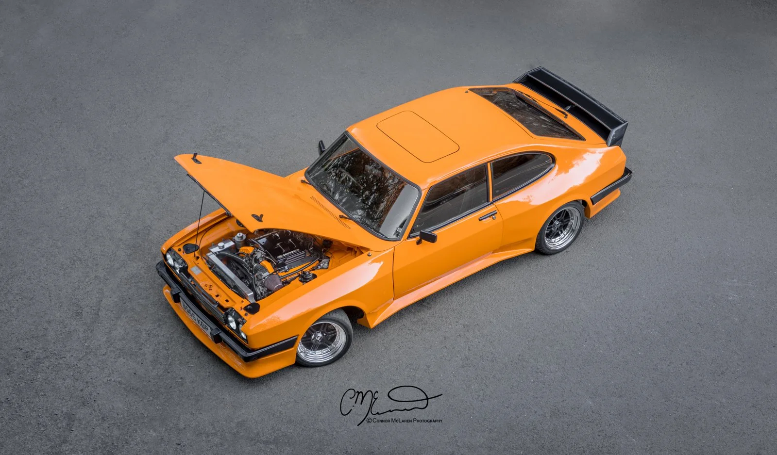 Ford Capri. An orange vintage sports car with its hood open, parked on a gray asphalt surface, showing the engine.