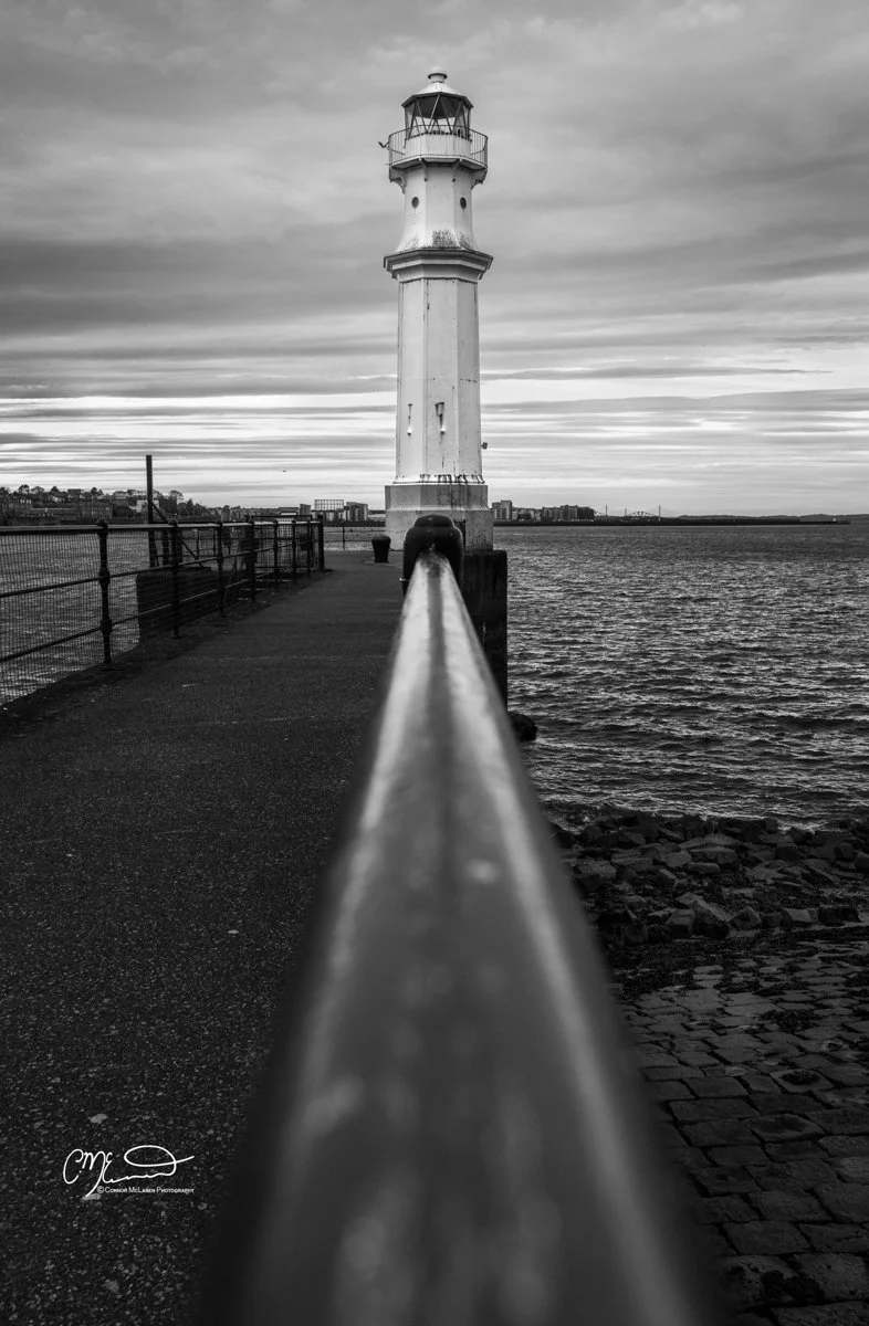 Scotland, Edinburgh. A black and white photo of a lighthouse at the end of a pier, with a metal railing along the walkway and a body of water on the right side, city buildings visible in the distance.