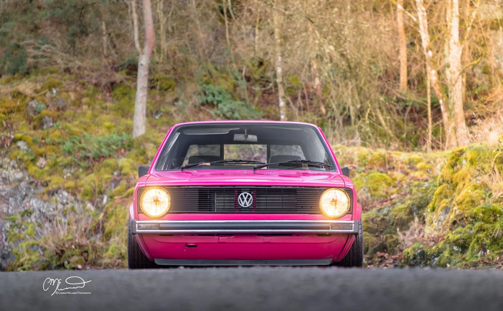 Front view of a pink vintage Volkswagen car on a road, with a forest background.