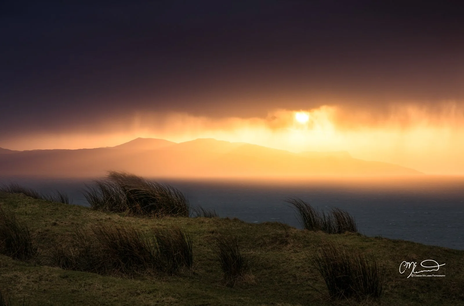 Scotland, Isle of Skye, Elgol. A landscape with grassy hills in the foreground, ocean in the middle ground, and a dramatic sky with clouds and sunlight breaking through in the background.