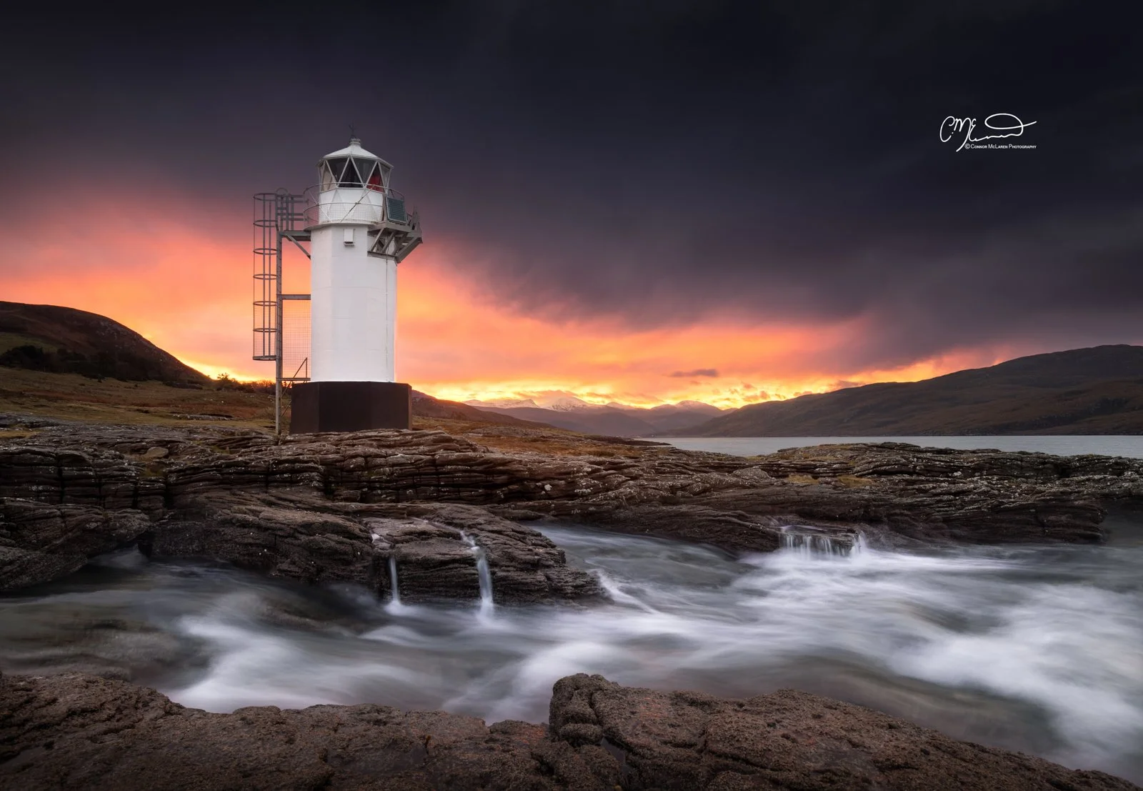Scotland, north coast 500, NC500, Rhue Lighthouse. A lighthouse on rocky terrain near water at sunset, with clouds and mountains in the background.