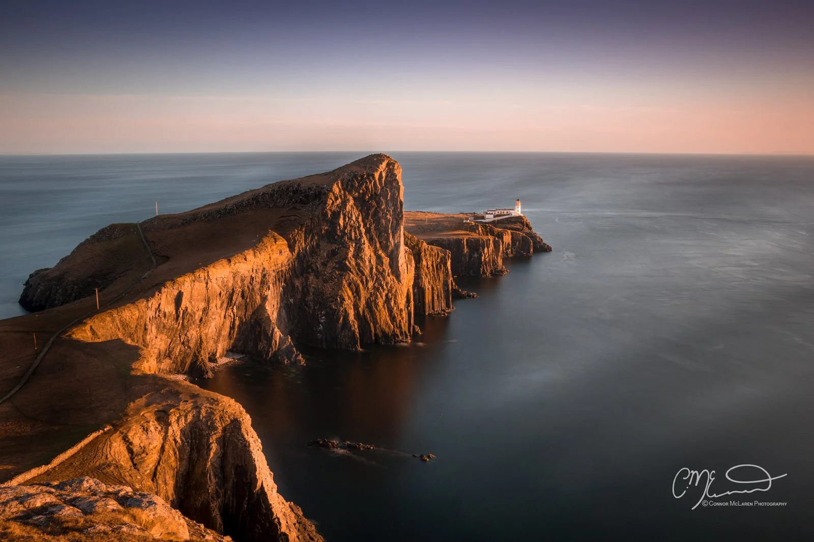 Scotland, Isle of Skye, neist point. Aerial view of a rugged coastal cliff with a lighthouse at the tip, illuminated by the warm light of sunset, with calm ocean water surrounding it.