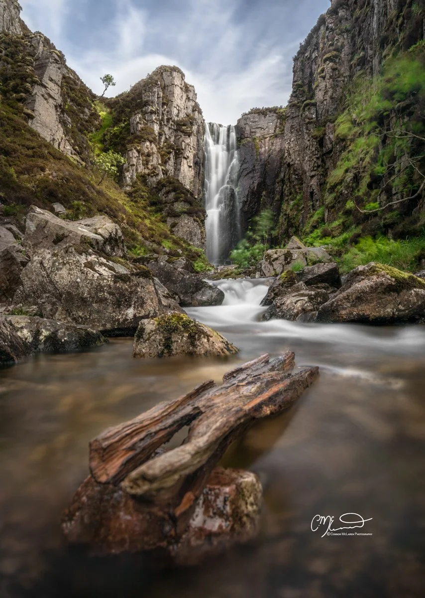 Scotland, north coast 500, NC500, Assynt. Wailing Widow Falls. Waterfall flowing between rocky cliffs with lush greenery, fallen logs in the stream, and a cloudy sky.