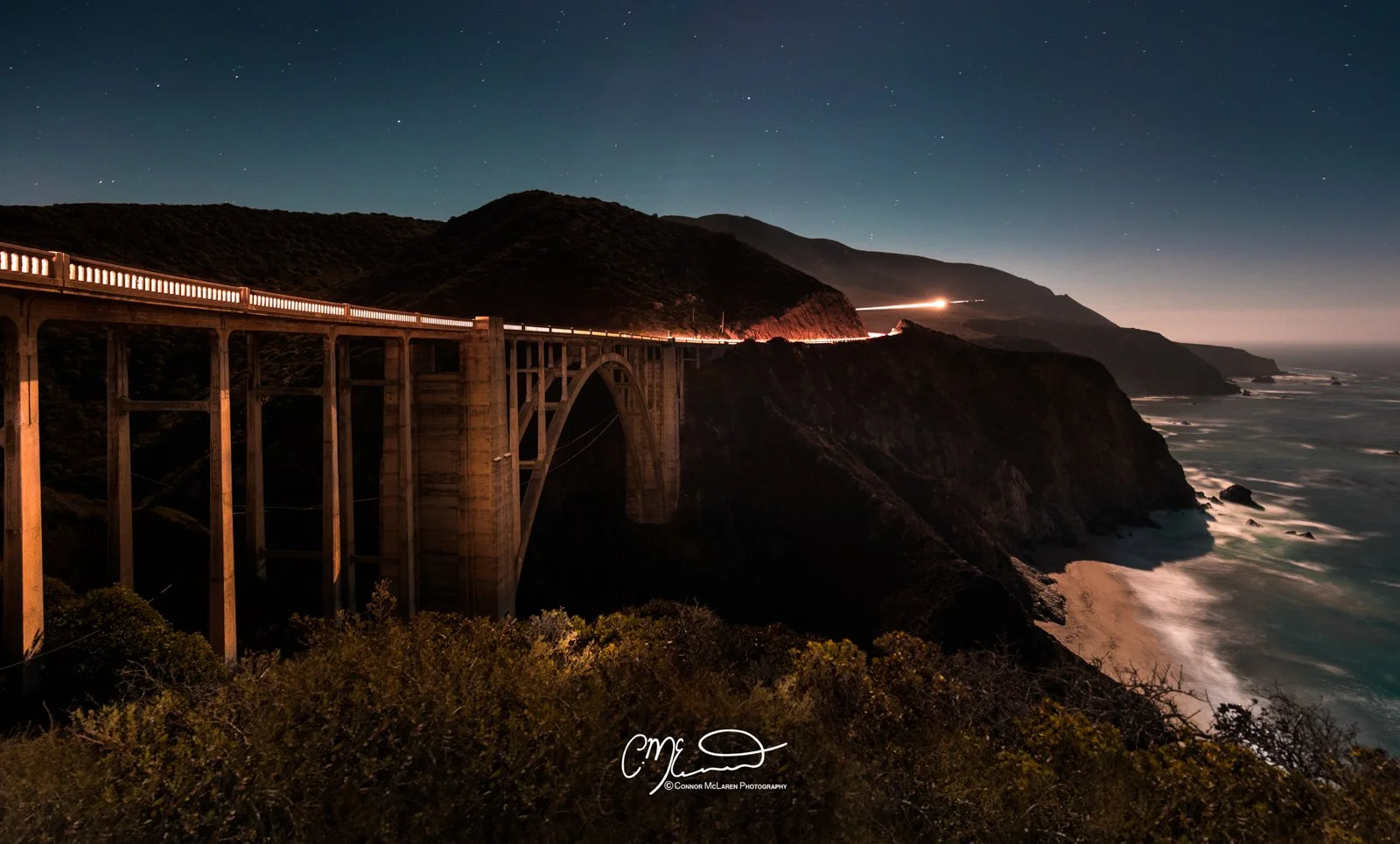 Big Sur bridge, California, United States of America, USA. Nighttime coastal scene with a lit-up bridge over cliffs, starry sky, ocean waves, and distant hills.