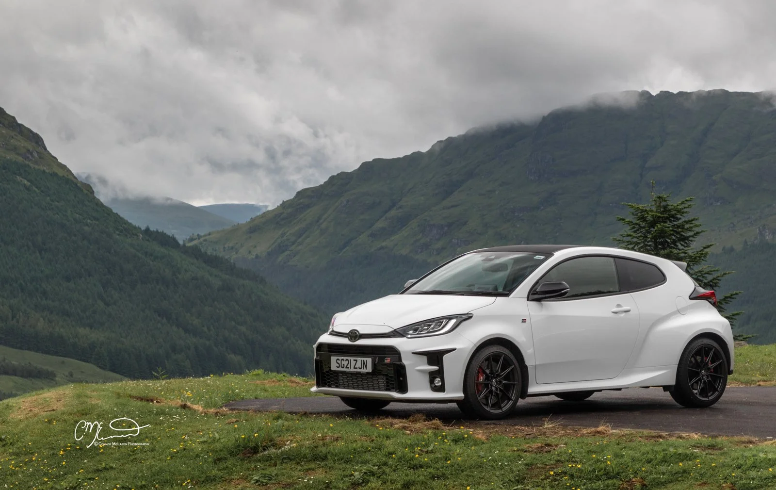 White sporty Toyota Yaris GR hatchback car parked on grass and blacktop with green mountains and cloudy sky in the background.