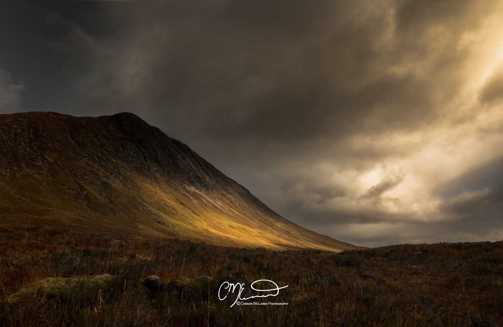 Glen Coe and Glen Etive, Western Highlands, Scotland. Landscape of a mountain under dark, cloudy sky with a patch of sunlight illuminating the hillside.