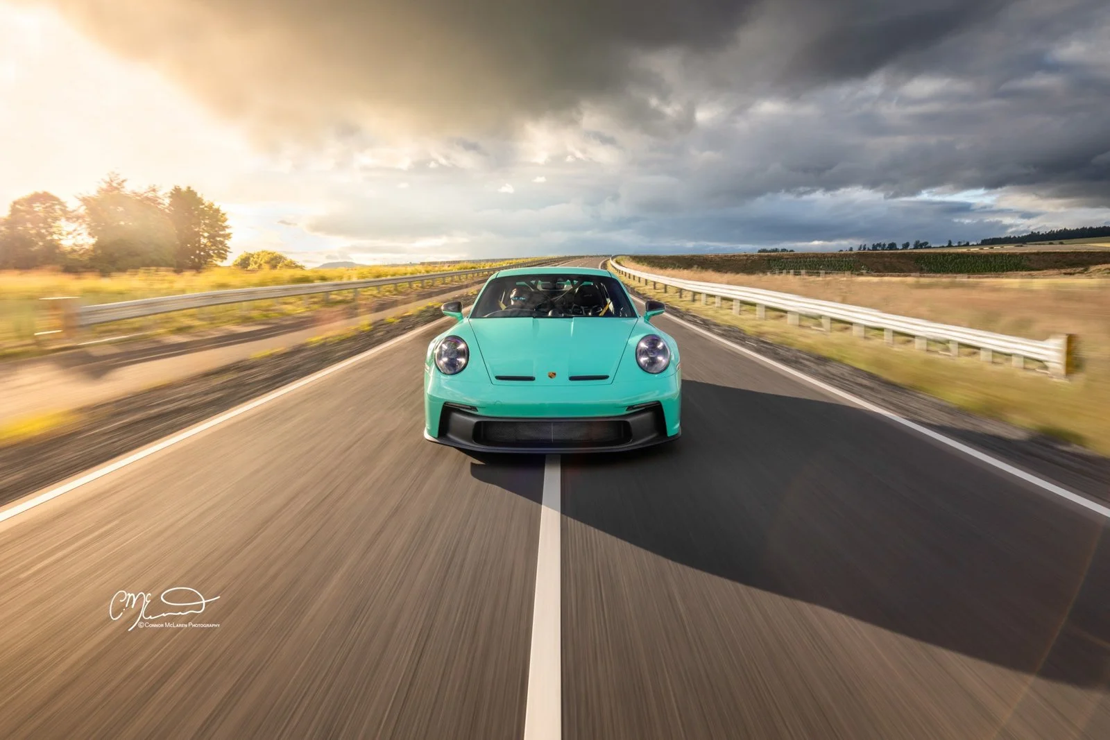 A Mint Porsche 911 GT3 992 sports car driving on an open highway with guardrails, with a cloudy sky and the sun setting in the background.