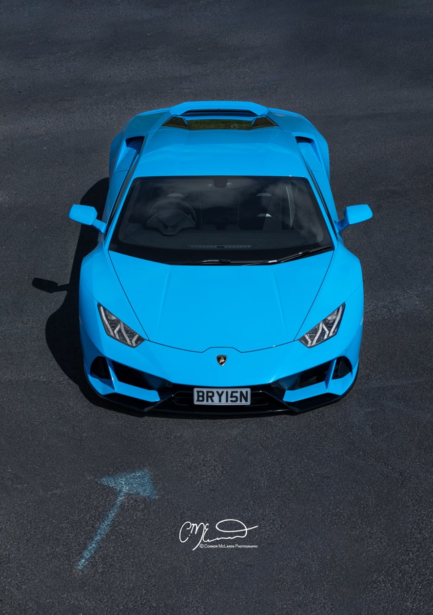 Blue Lamborghini huracan evo sports car photographed from above on a dark asphalt surface with a faint blue arrow pointing left painted on the ground.