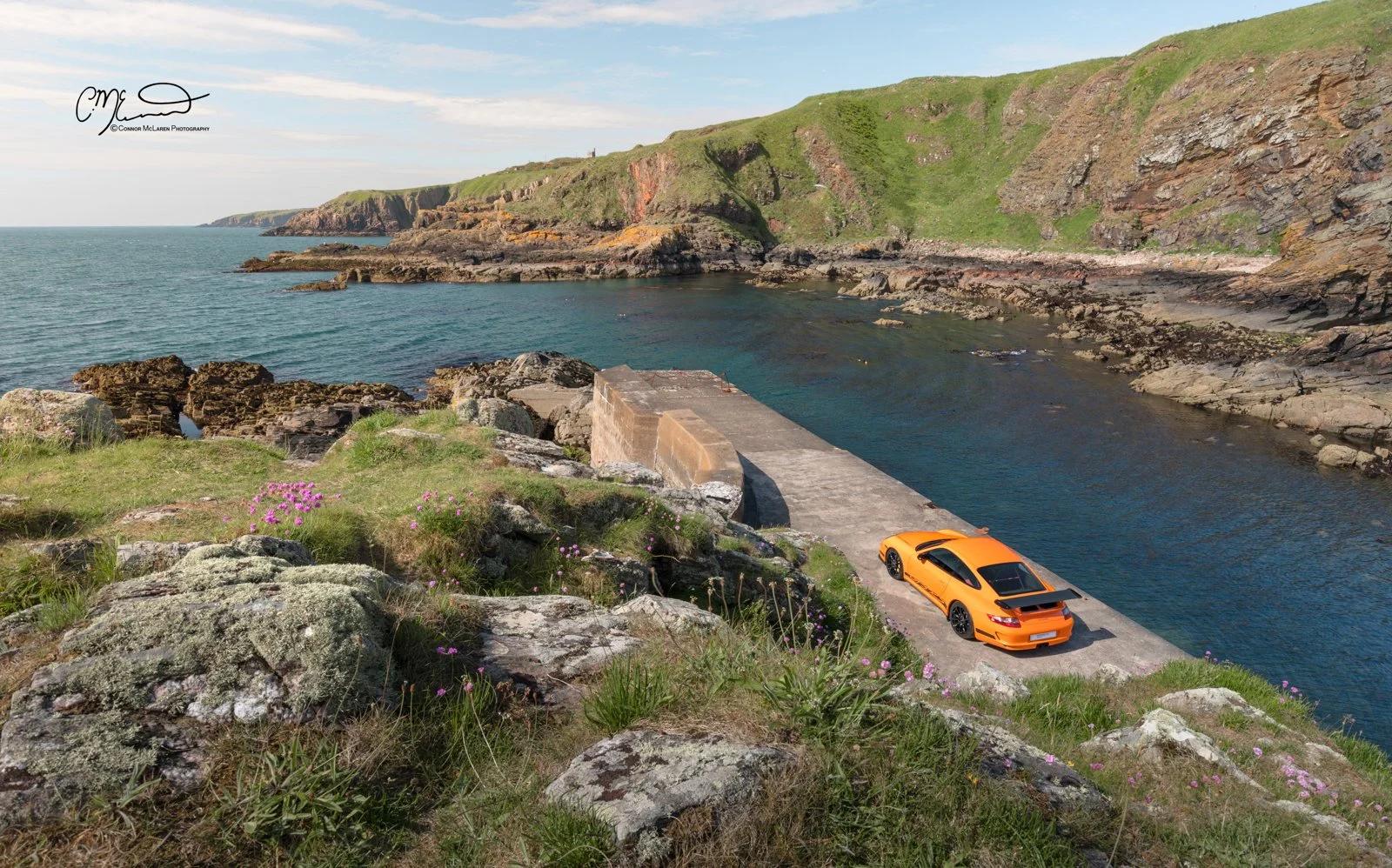 Porsche 911 GT3 RS, 997. An orange sports car parked on a concrete slipway along a rocky coastline with green cliffs in the background, pink wildflowers in the foreground, under a partly cloudy sky.