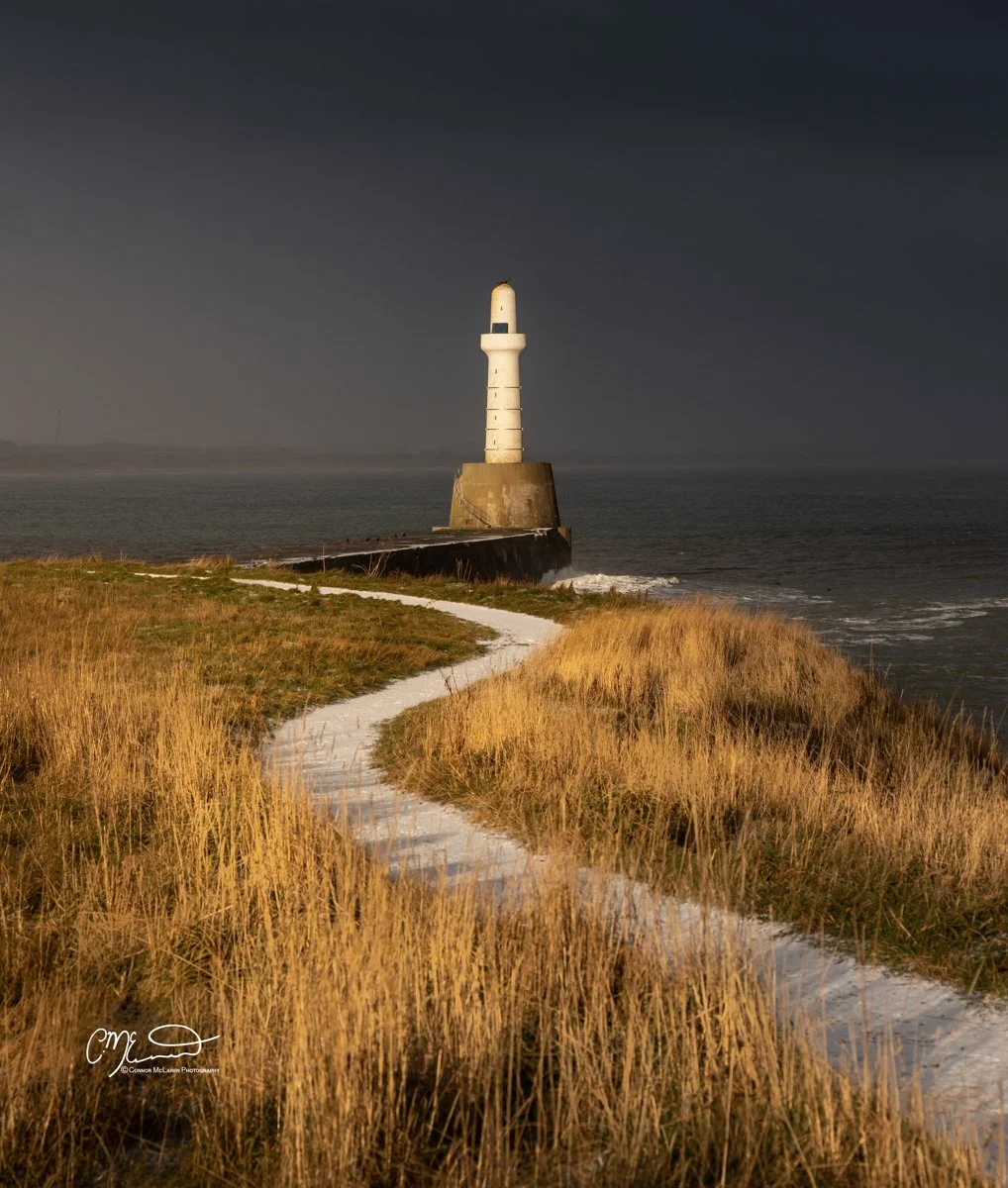 Aberdeen Lighthouse, North East Scotland. A lighthouse on the coast during sunset, with a path leading through tall grasses towards it, and dark sky in the background.