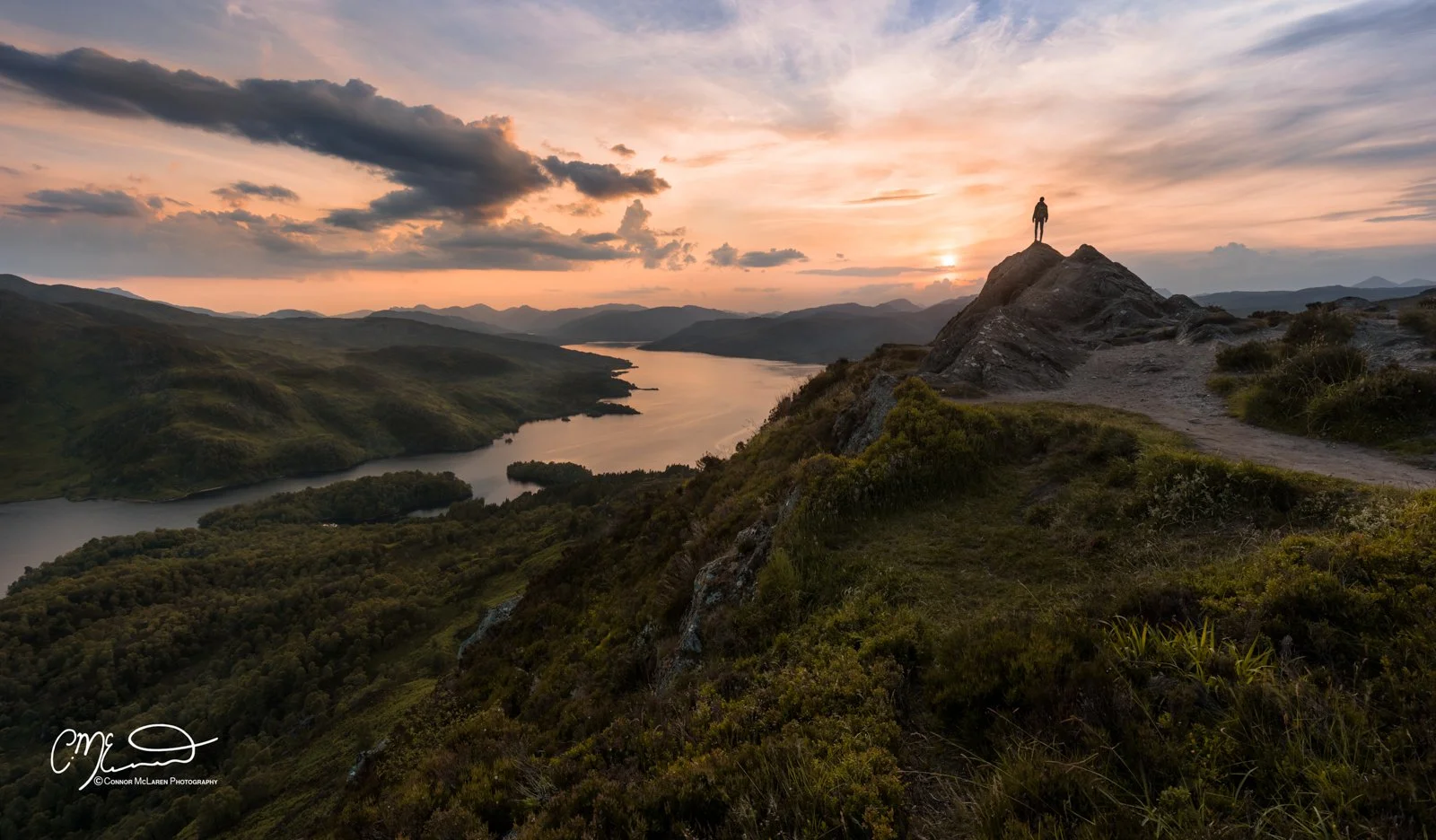 Scottish Lowlands, Scotland, ben ann. A person standing on a rocky outcrop overlooking a winding river valley during sunset, with mountains in the distance and a partly cloudy sky.