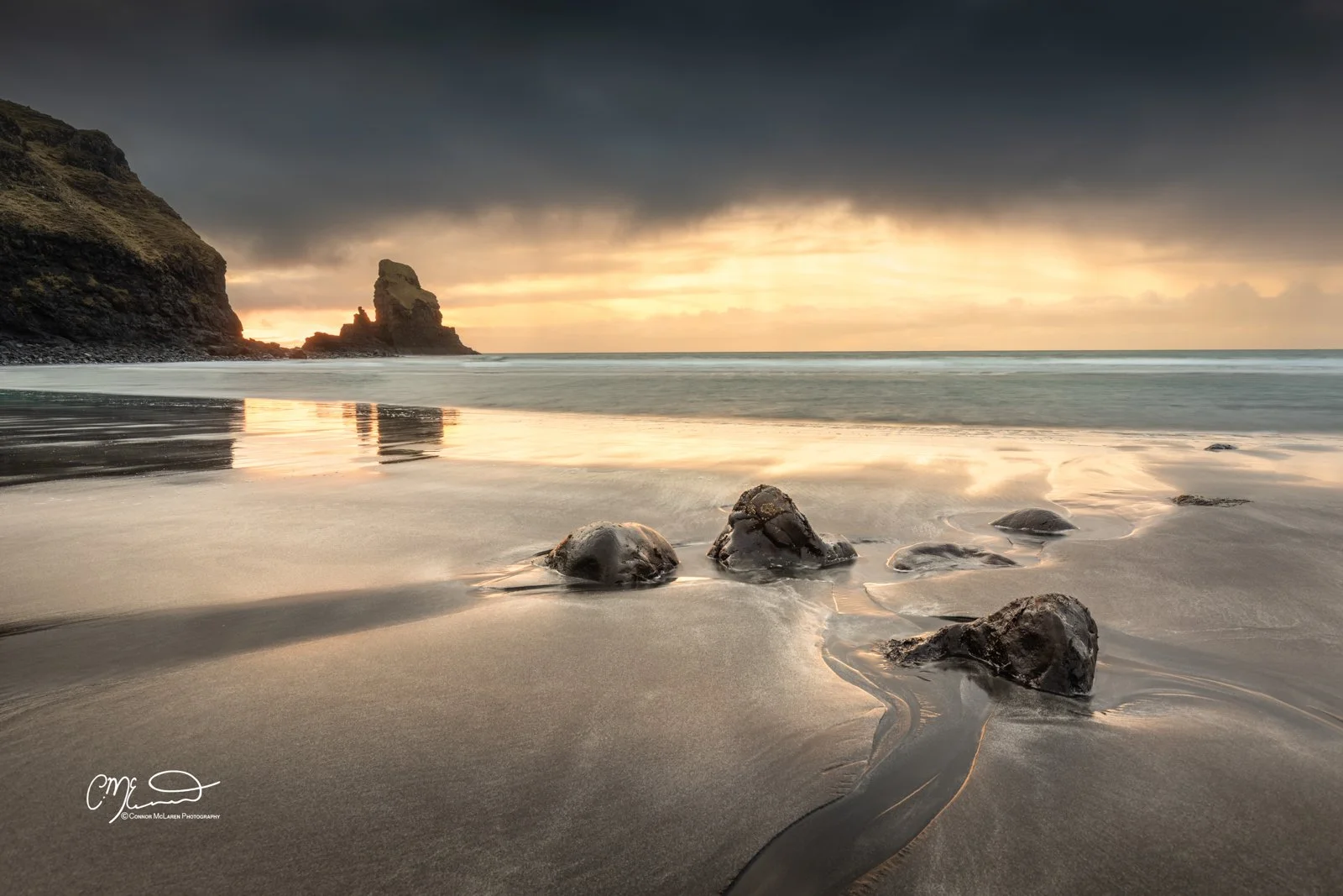Bucket list locations, Scotland, Isle of Skye, Talisker bay. Sunset over a beach with rocks in the sand and a large rock formation in the distance, with dark clouds overhead and reflections on the wet sand.
