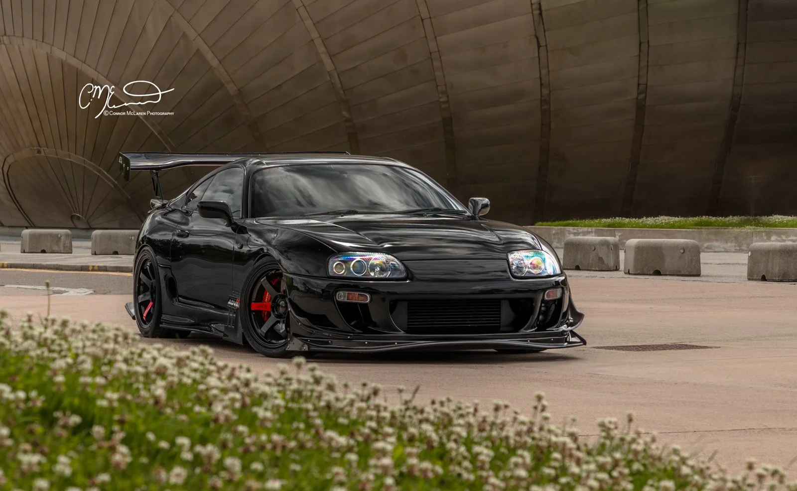 Custom Modified Toyota Supra 2JZ. A black sports car with a large rear wing and red brake calipers parked on a paved area near concrete barriers, with a large, curved modern architectural structure in the background.