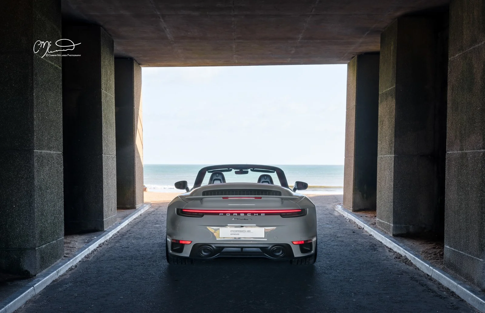 A silver Porsche 911 Turbo S convertible parked under a concrete tunnel with an ocean view in the background.