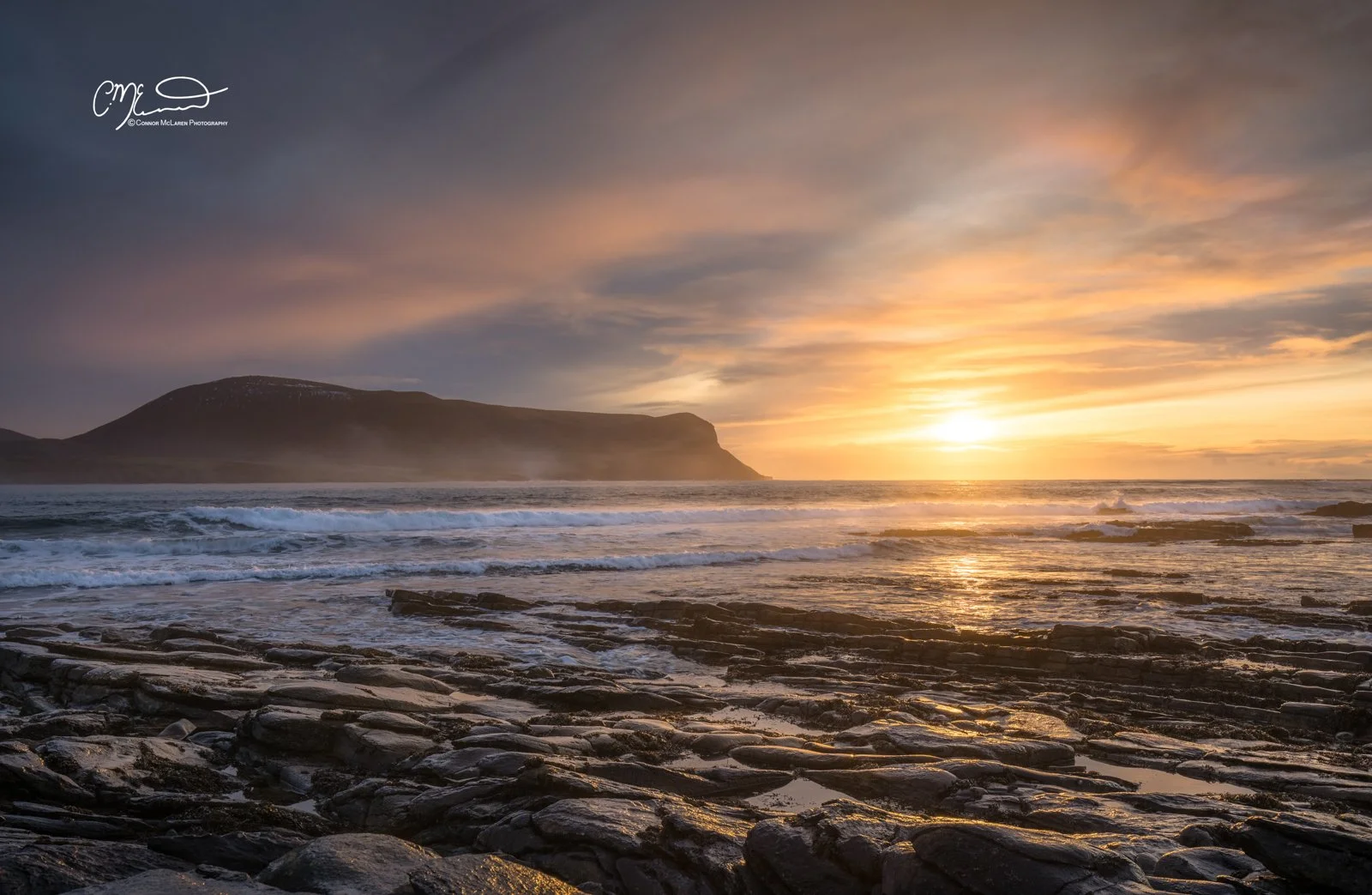 Orkney, Scotland. Sunset over the ocean with rocky shoreline and hills in the distance.
