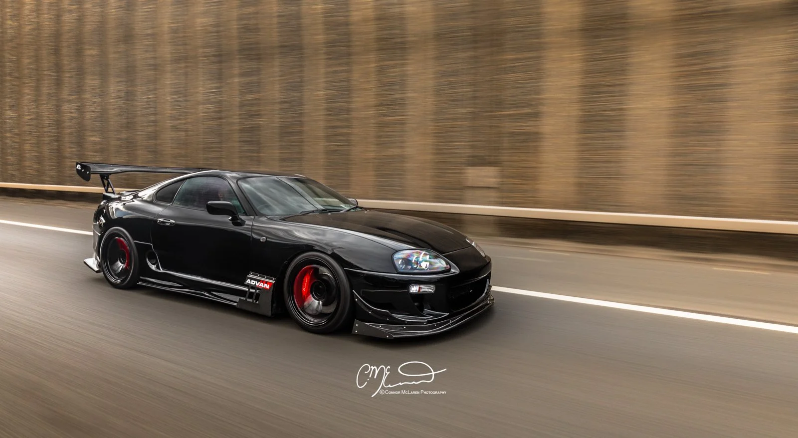 Custom Modified Toyota Supra 2JZ. Black sports car with red accents driving on highway with brown wooden guardrail and background