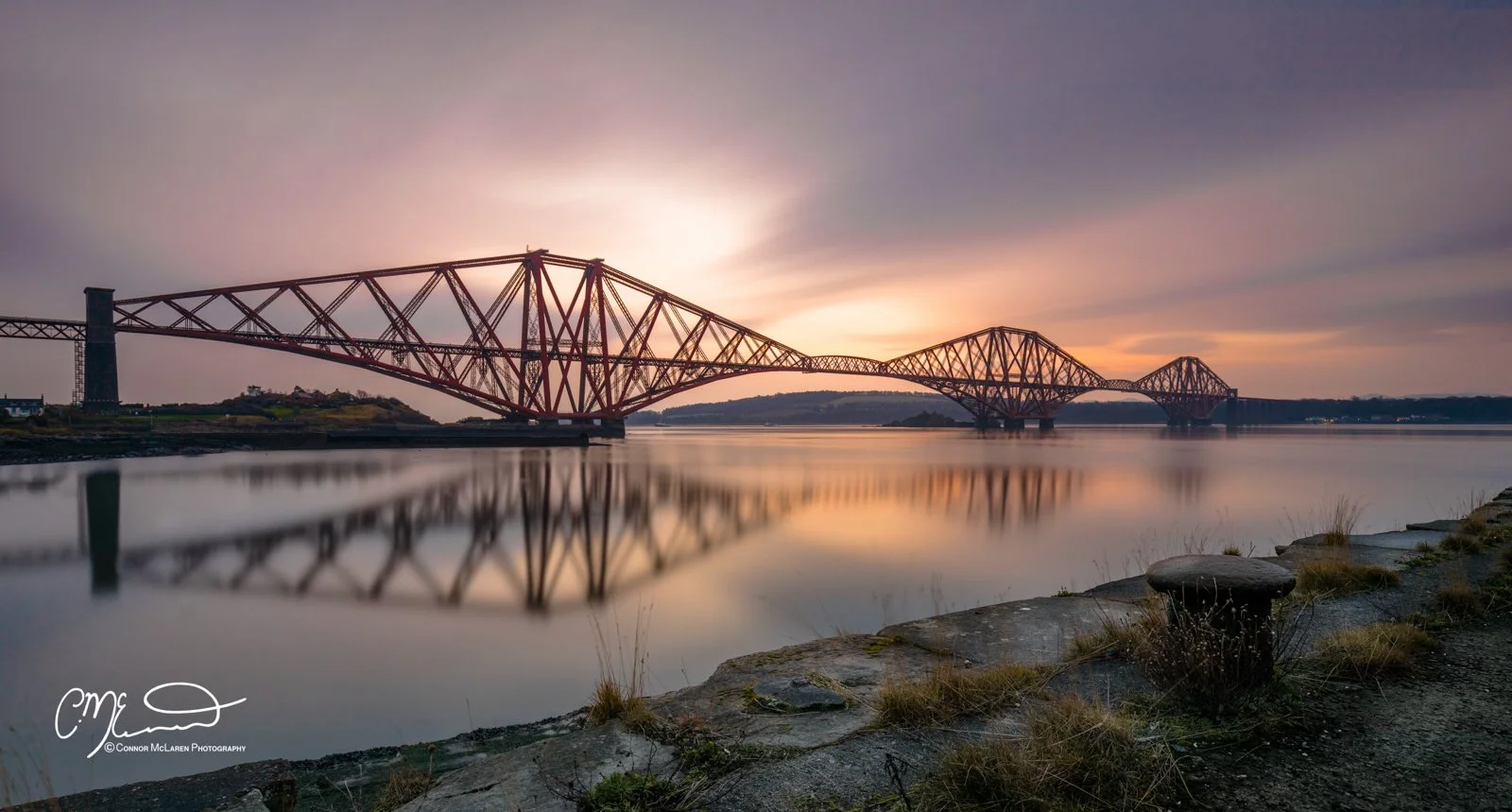 Scottish Lowlands, Scotland, Forth bridge Edinburgh. A large red bridge stretches across a calm river at sunset, with its reflection visible in the water and a rocky shoreline in the foreground.