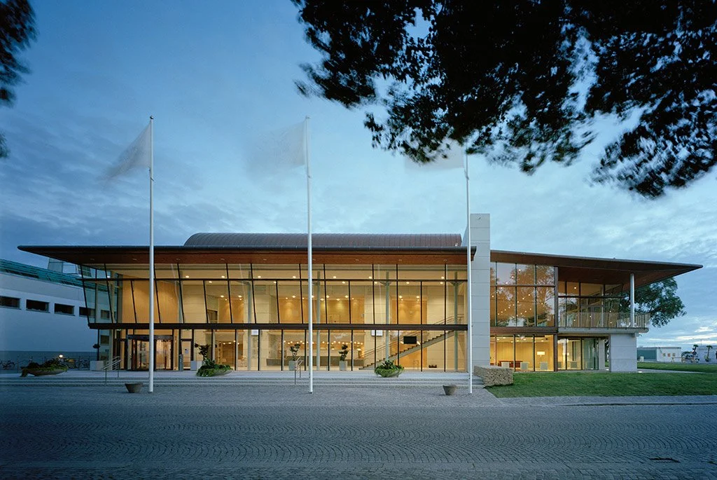Ett modernt kontors- eller bibliotekshus med stora glasfönster och tre flaggstänger framför. Byggnaden är upplyst inomhus och omges av en öppen plats och gräs.